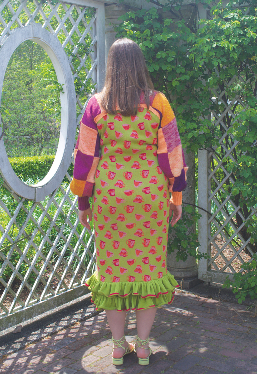 Back view photo of a model wearing a green, teacup-motif dress with a ruffle hem and a purple-and-orange patchwork turtleneck shrug. She is standing next to an oval-shaped window.