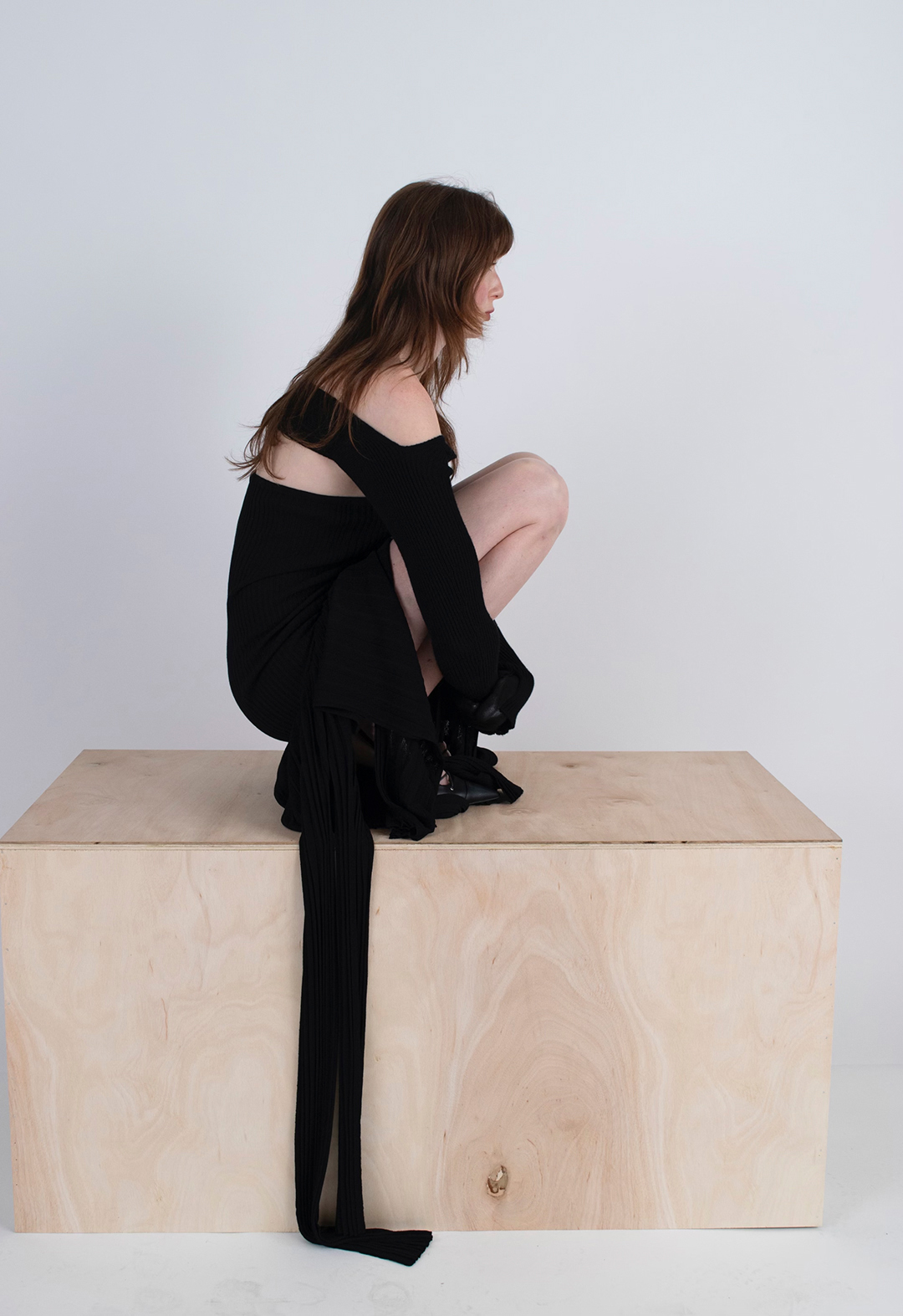 Model sitting down on a piece of wooden block. The model is wearing a black sculpted dress with variegated pleated panels.