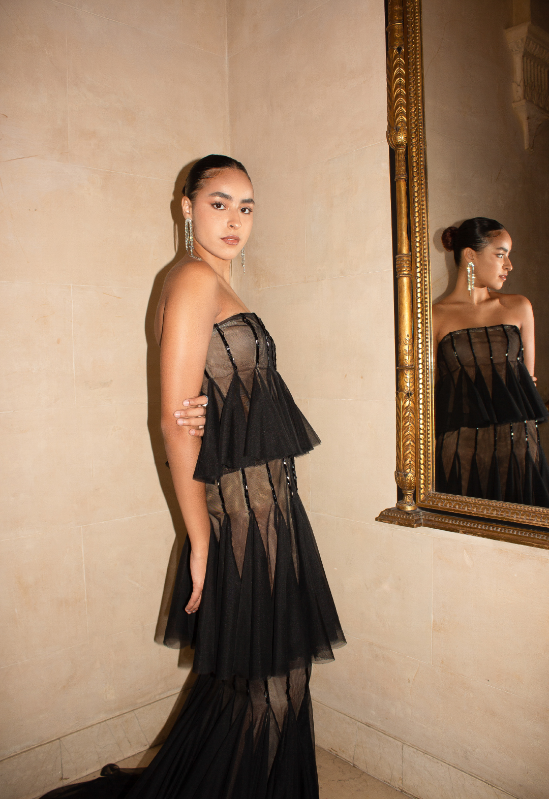This is the side view of the embellished black mesh gown. In the image, the model is standing in front of a gold-framed mirror and looking into the camera. Her image is reflected in the mirror. The image is giving a very royal vibe. The look is complemented by a pair of long rhinestone earrings.