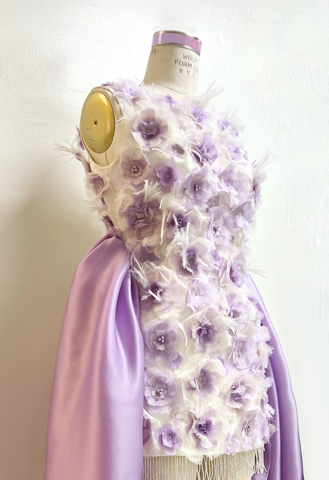 This dress is made of silk satin, organza, and chiffon. Detailed photo of a white satin mini dress with lilac embellished chiffon and organza flowers on a dress form. Flowers made of chiffon and organza that are different shades of lilac. Iridescent beads and swarovski crystals can be seen on the center of each flower.