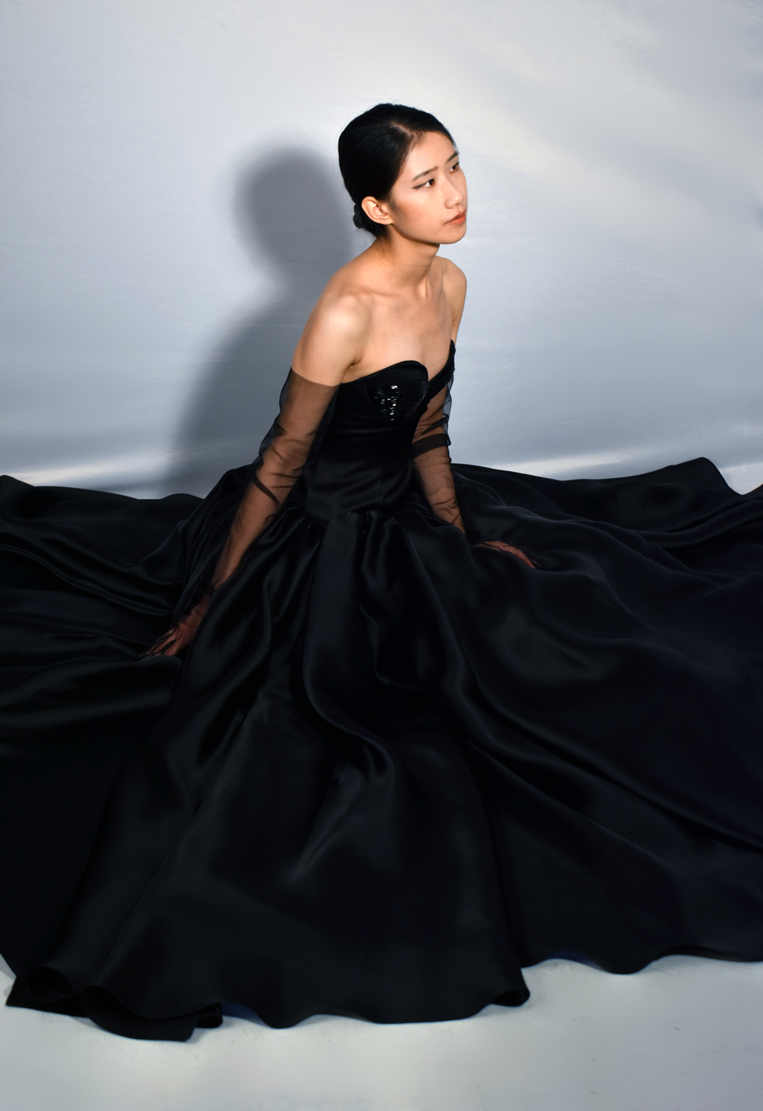 This black gown is made from 100 percent silk. The photo shows a woman sitting in a three-quarter view, wearing a black silk gown with beading covering three-quarters of the bodice cups. The floor-length gown has a drop-waist duchesse satin bodice with cone-like satin-faced organza skirt panels gathered along the drop-waist, spread onto the floor as she sits. The model is wearing black tulle gloves that cover three-quarters of her arms. Her head is turned away, looking up to the side. The background is white.