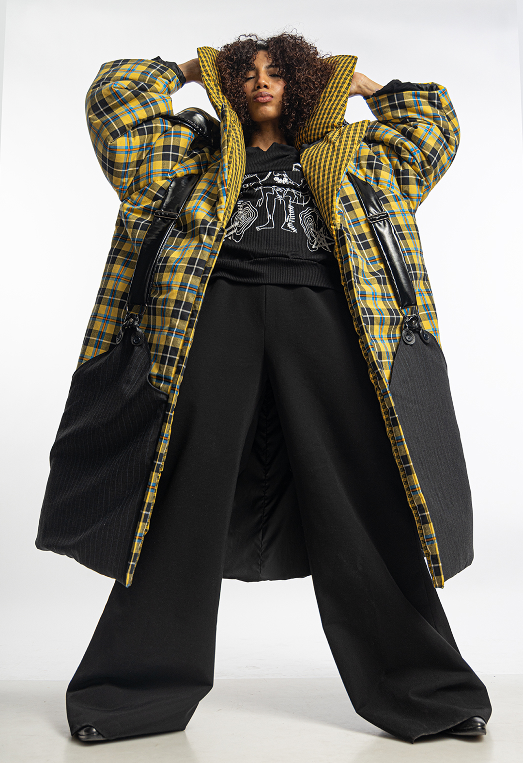 A model is wearing a yellow plaid padded coat with oversized padding, a wide yellow tartan collar, and huge pinstripe pockets, over a sweatshirt and black flare pants