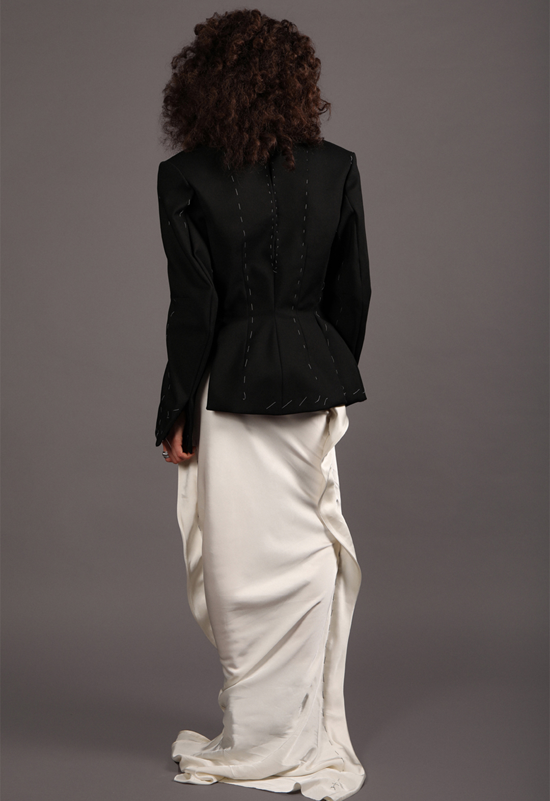 Model wearing a black tailored jacket with basting details, paired with a silky bias-cut ivory skirt with basting details, viewed from a back perspective.