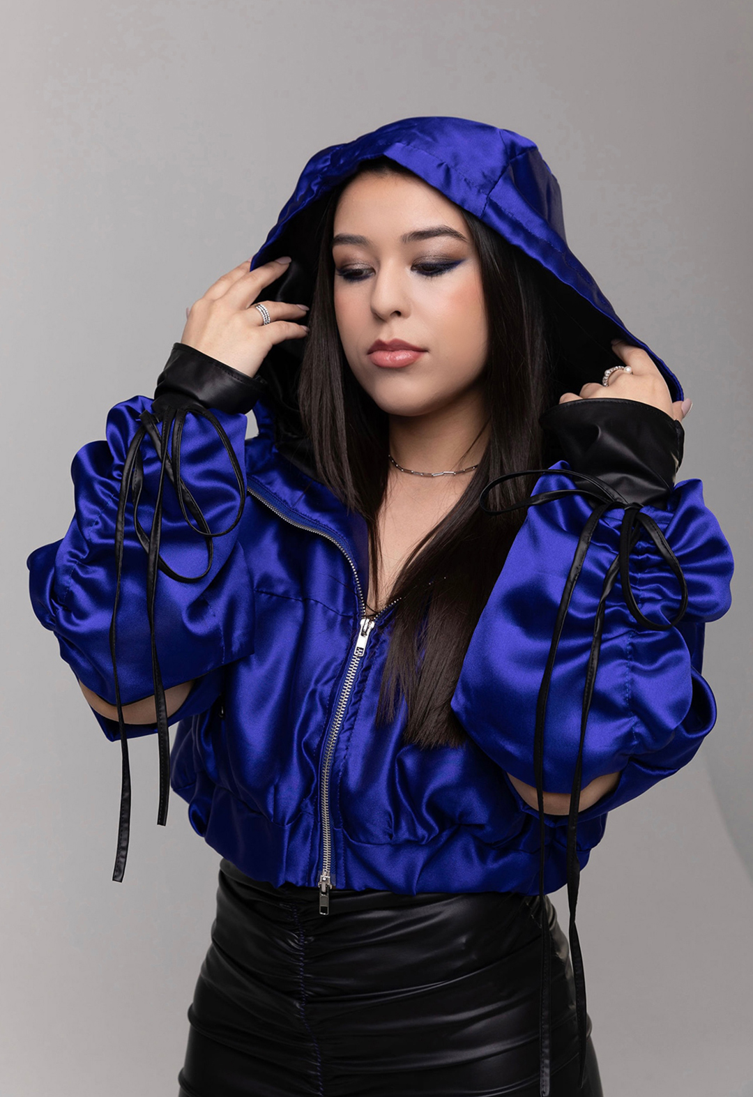 This is a close-up photo of a model wearing a royal blue jacket. She has the hood on and she has her hands on the hood. There are black drawstrings at the cuff of the jacket. The background is gray. 