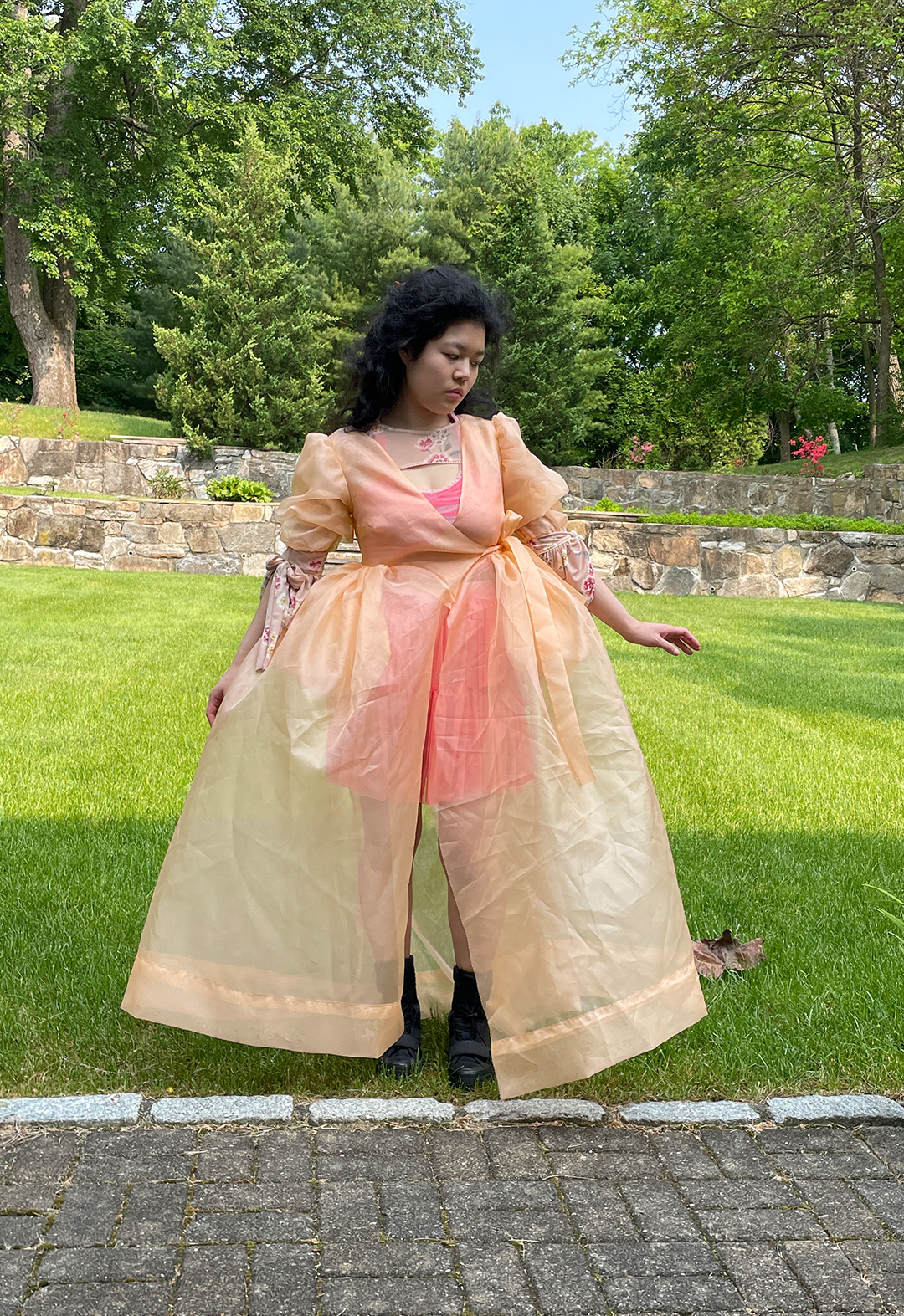 Front view of a model standing in a garden and wearing a translucent peach gown. The model is wearing a tiered skirt under the gown.
