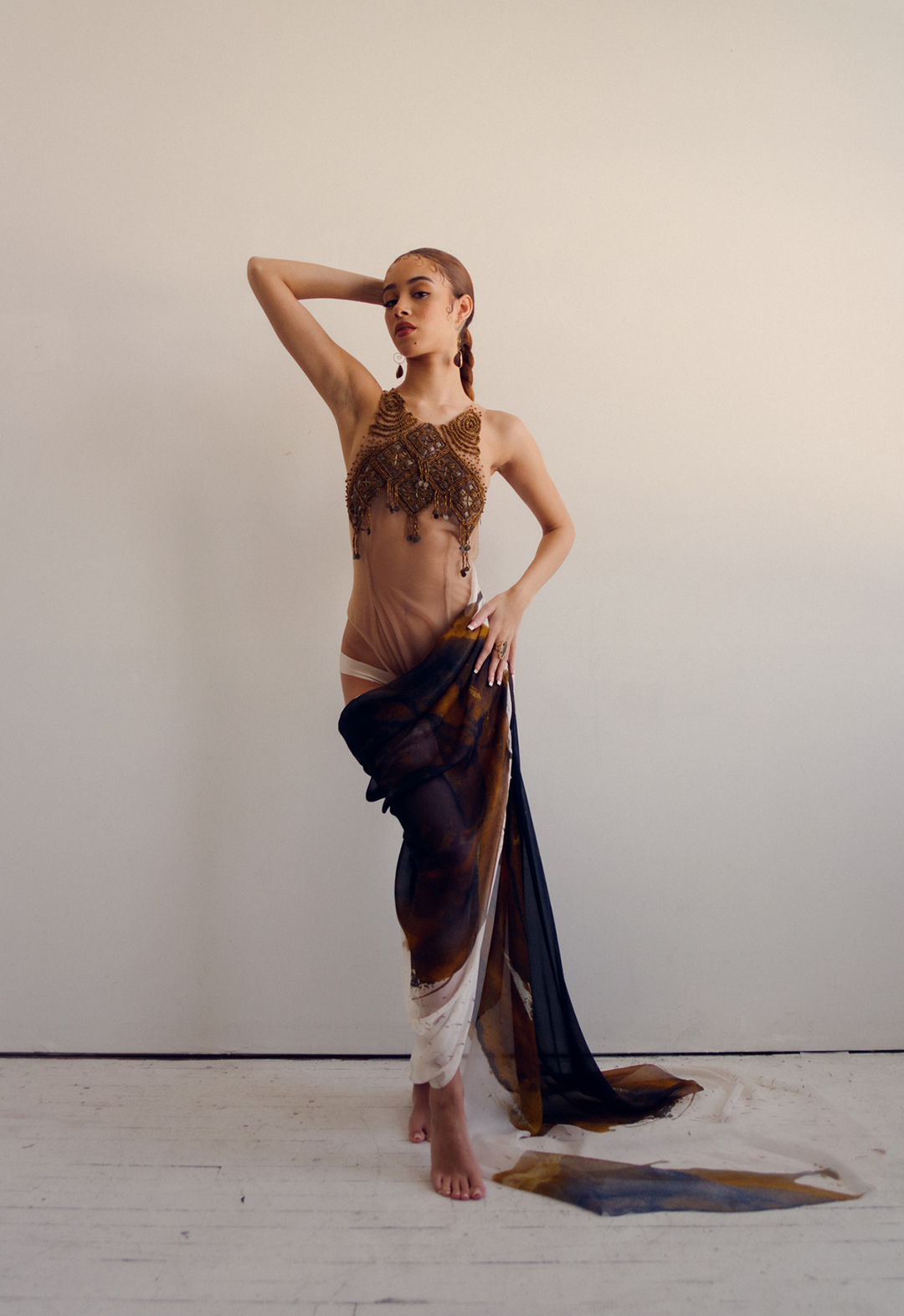 Front view of a model wearing a brown and white evening gown, made of mesh and hand beading. One of the model's hands is on her hip and the other one is on the back of her head.