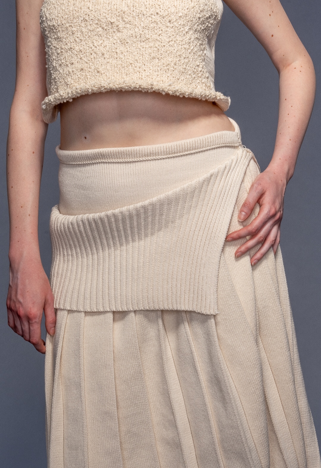 Model wears funnel neck top with misshaped intarsia and irregular pleated skirt in organic cotton. Her hair is tucked in the sweater and she is looking to the right.