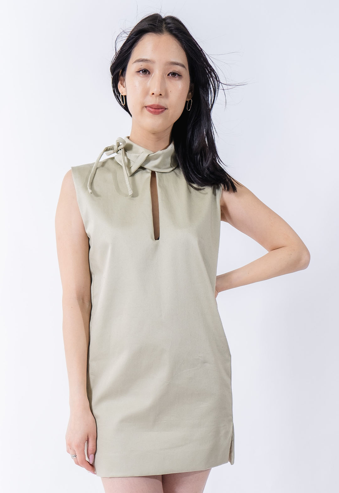 This pale, light-green mini dress has a skinny rectangular cutout from the neck to the bust. There is a stand collar attached to the neckline that pulls through itself via welt openings to tie into a bow at the side of the neck. The model is standing in front of a white background and looking straight into the camera with one hand on her hip.  