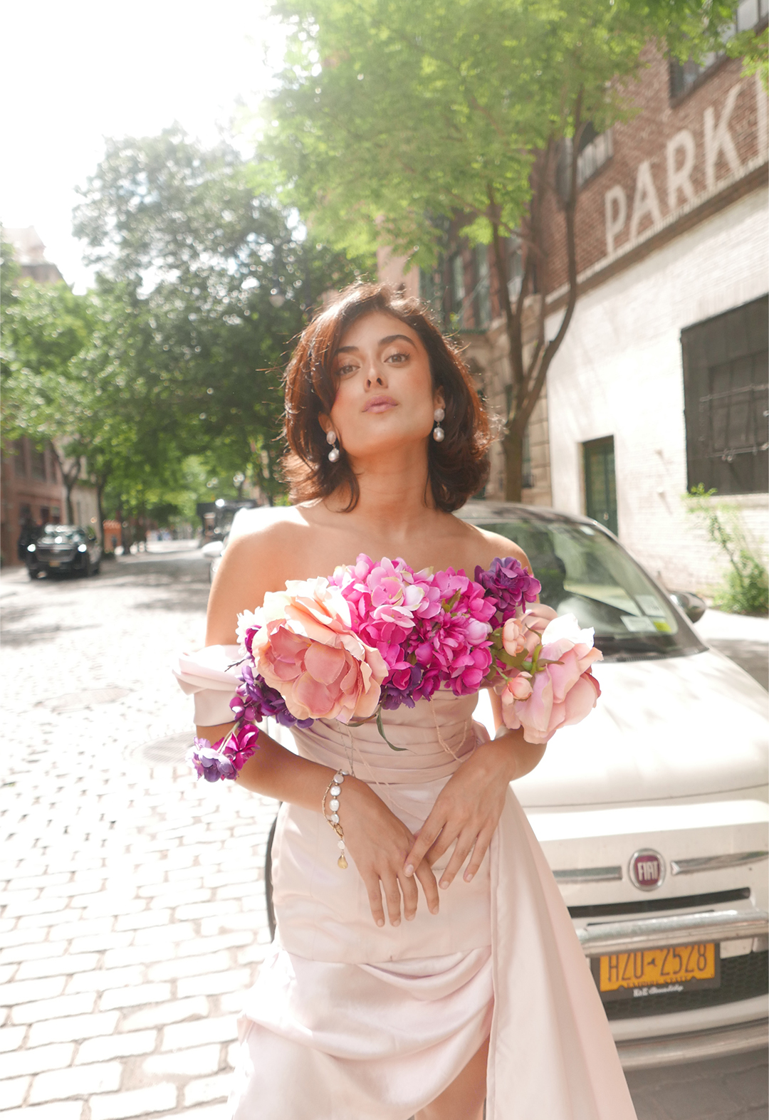 Photo of a model standing in the streets, wearing a baby pink off-shoulder dress with draped cowl and side train. There are many flowers on the neckline of the gown. The model has her elbows bent and both her hands in front of her, with just her fingers touching each other. 
