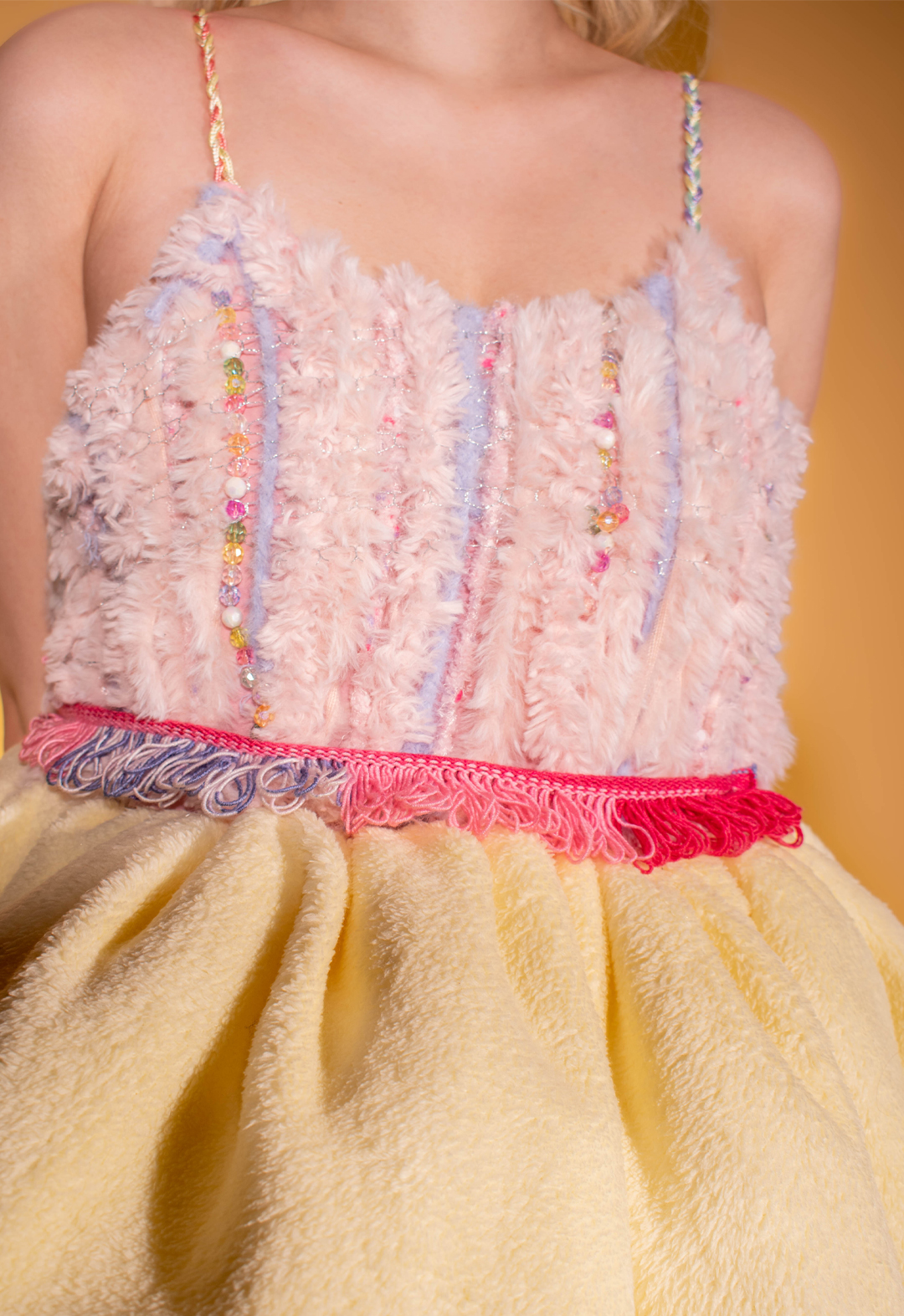 Photo of the detail of the bodice and belt of the garment. The stripes are braided into colorful stripes. The fabric of the bodice is a weaving manipulation with pink-and-blue furry yarns and colorful beading. The belt is knitted in pink yarns with tassels.