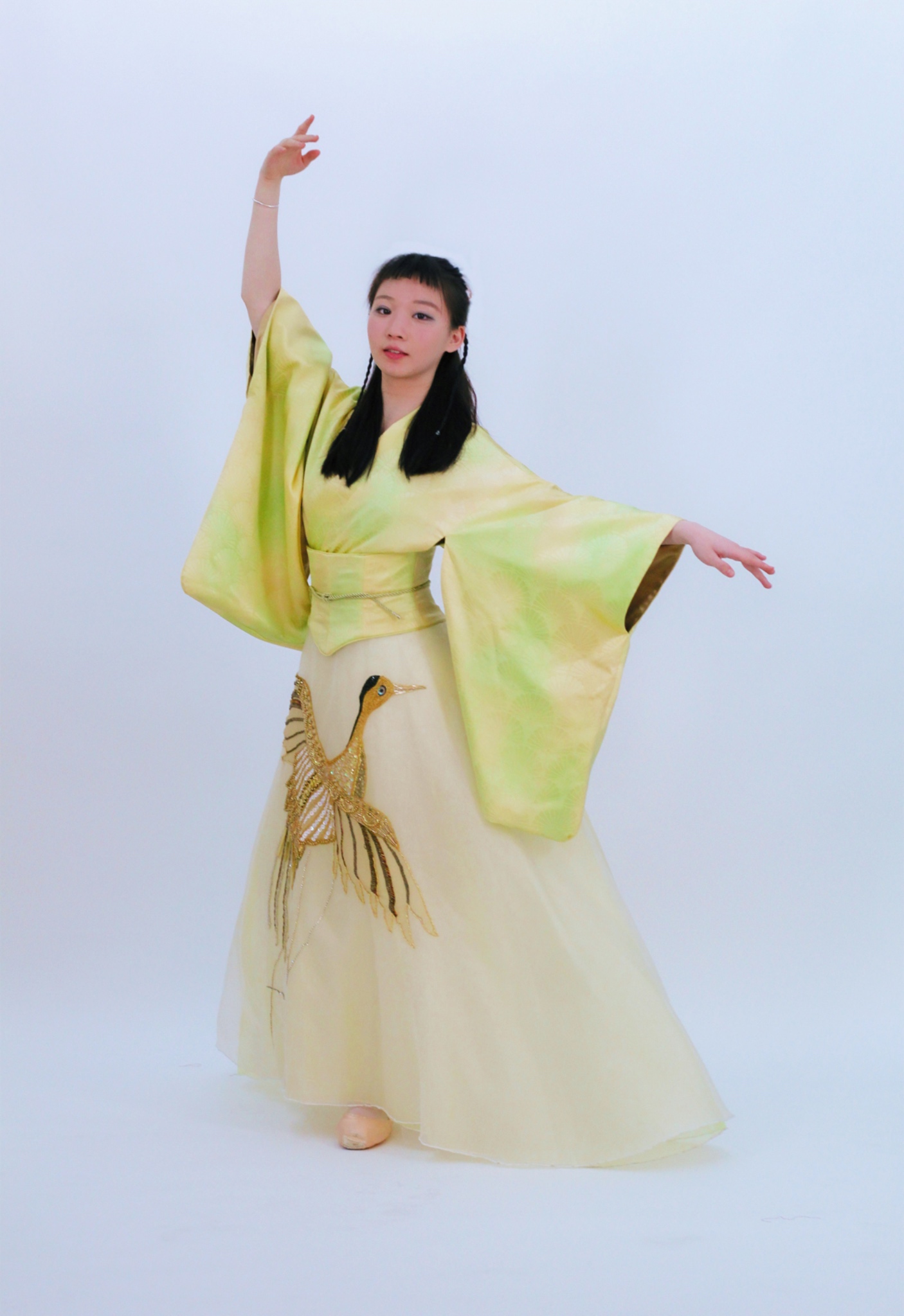 A full view of a ballerina in a yellow silk kimono-inspired dress. She is posing with one arm up and one arm to the side. 