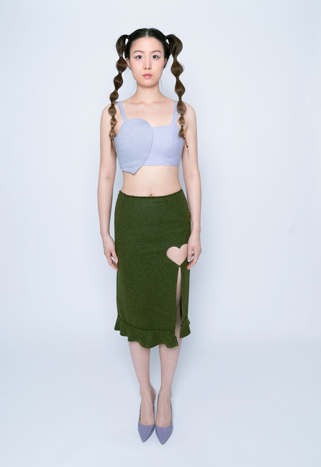 A three-quarter view of a model playing with her hair, which is styled in two bubble ponytails. There is a purple heart patch on the right side of the top, a heart-shaped cutout and split on the right side of the moss-colored skirt, with a ruffle at the hem. 