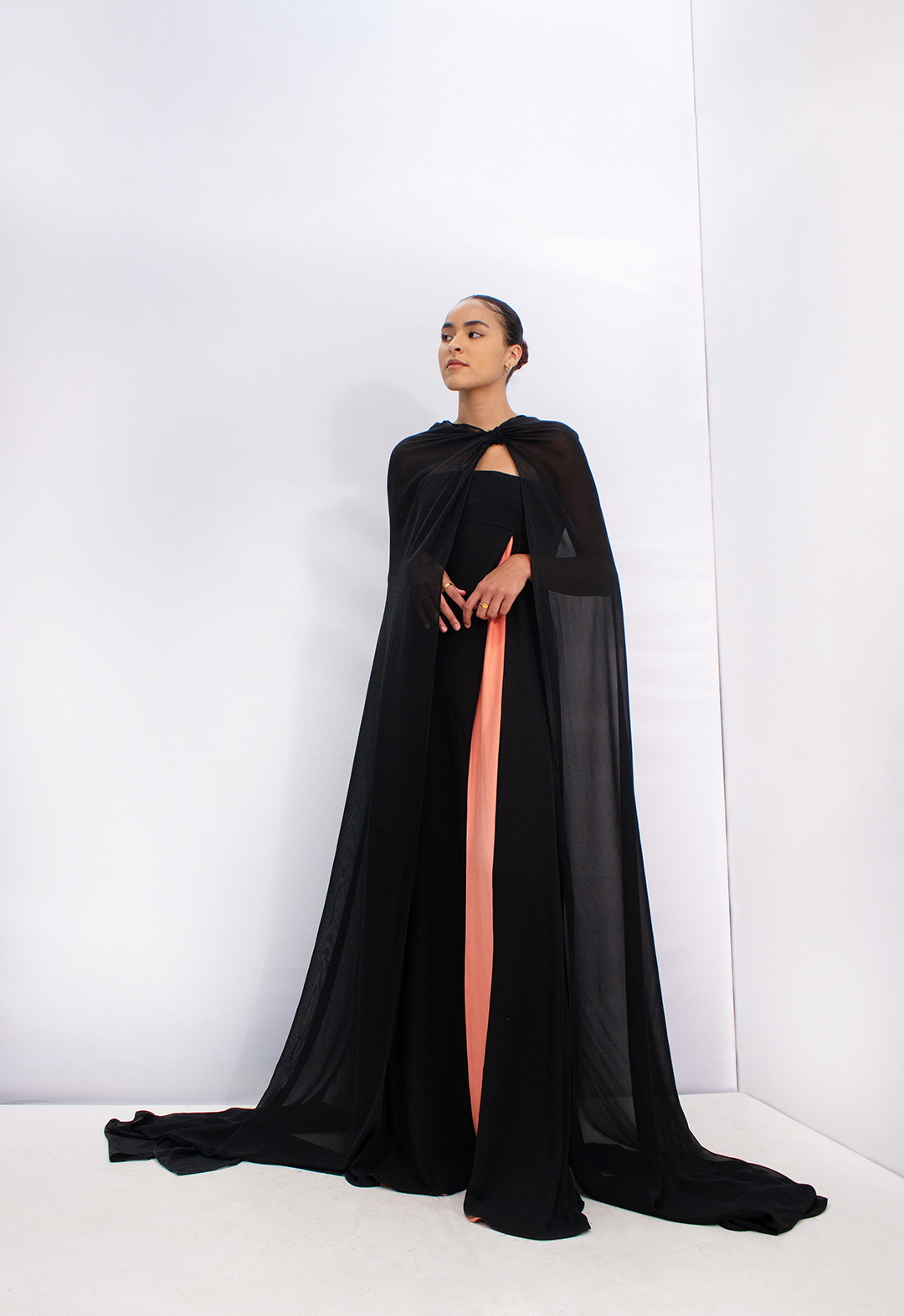 The front view of the dress with a cape. This black georgette dress has a front slit that has a coral charmeuse skirt underneath. The black chiffon cape has a knot on the left side of the wearer’s neck that drapes toward the back of the cape.