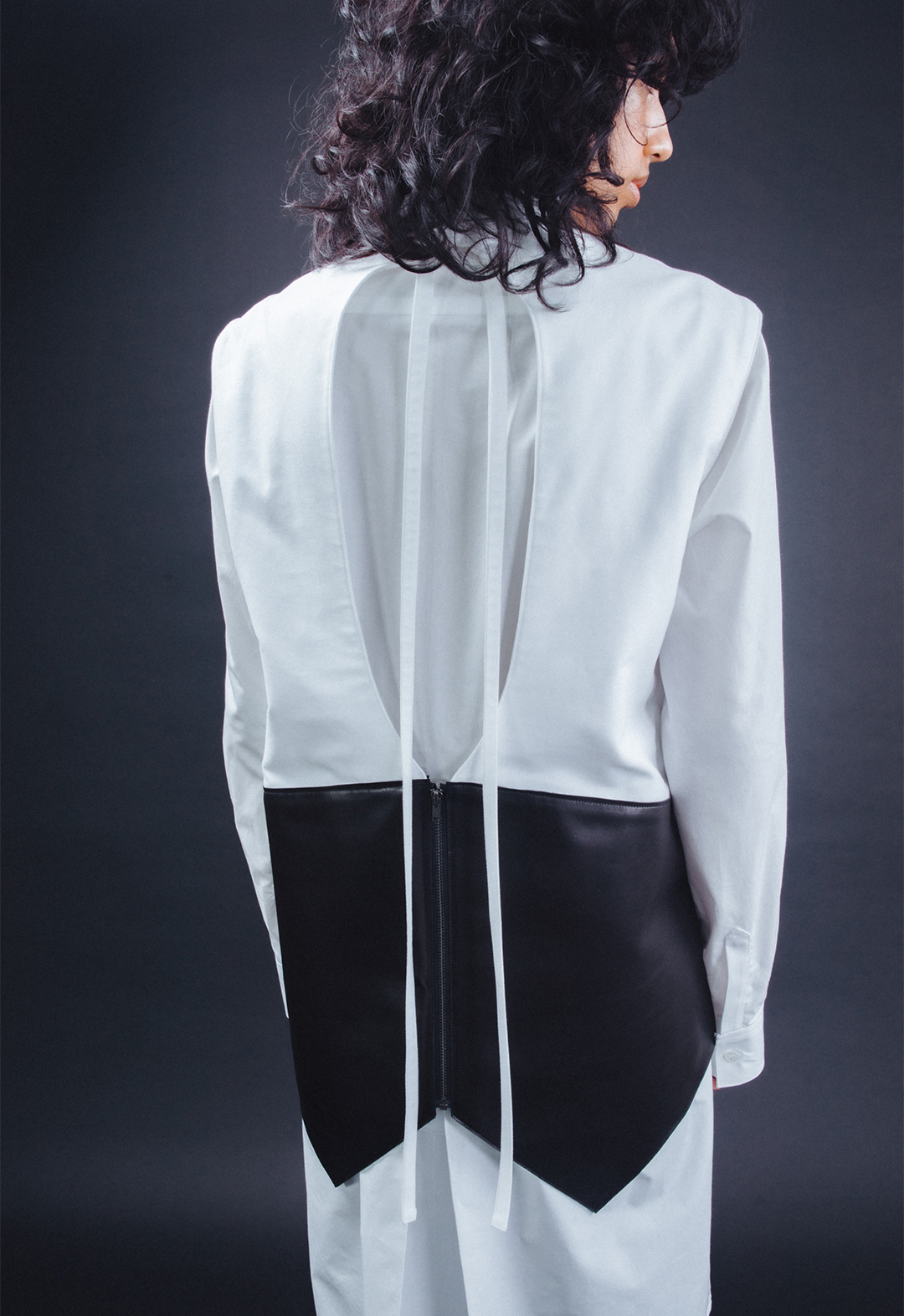 Photo of the model, Aria Puga, wearing a white cotton tunic shirt and a white cotton and black leather open-back top. The open-back top has a cut-out with a tie at the back neck and a zipper at the center back. The model is turned with their back to the camera and their head is turned to the side. The background is black.