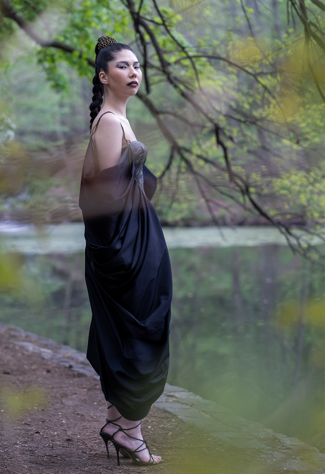 A woman in a black-and-gold dress stands with poise and elegance. Her dress exudes sophistication and elegance, with the flowing fabric draping gracefully around her legs emphasizing her confident demeanor. The surrounding environment features a rocky terrain in the forest. This image exudes an air of mystery and intrigue, leaving much to the imagination regarding the woman’s fierce attitude.