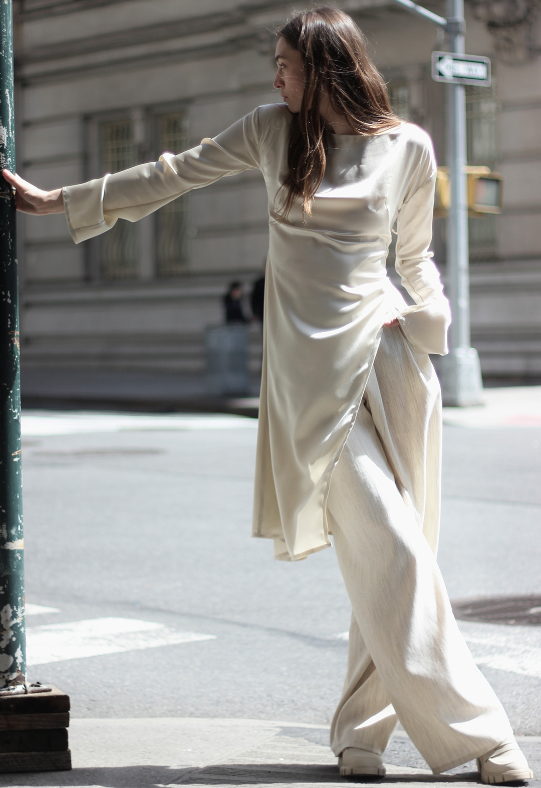 The model is looking to the side with one arm on a pole and her other hand in her pants pocket.  She has one leg crossed over the other. She is wearing a cream silk tunic with a high neck and a side cutaway to highlight her cream pleated wide-legged pants.