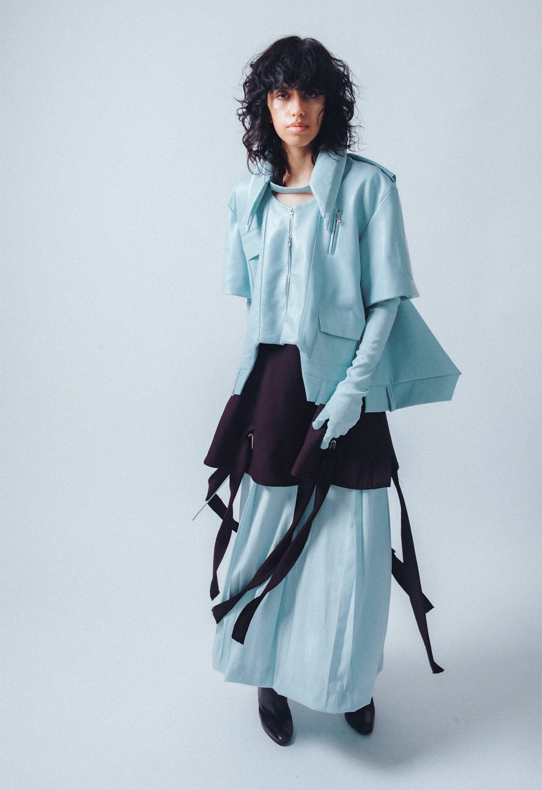 Front view of look 2, showing a baby-blue, double-collar leather shirt jacket with trapunto details and a back opening, a baby-blue double-pleated skirt, and a dark-purple grommet apron skirt.