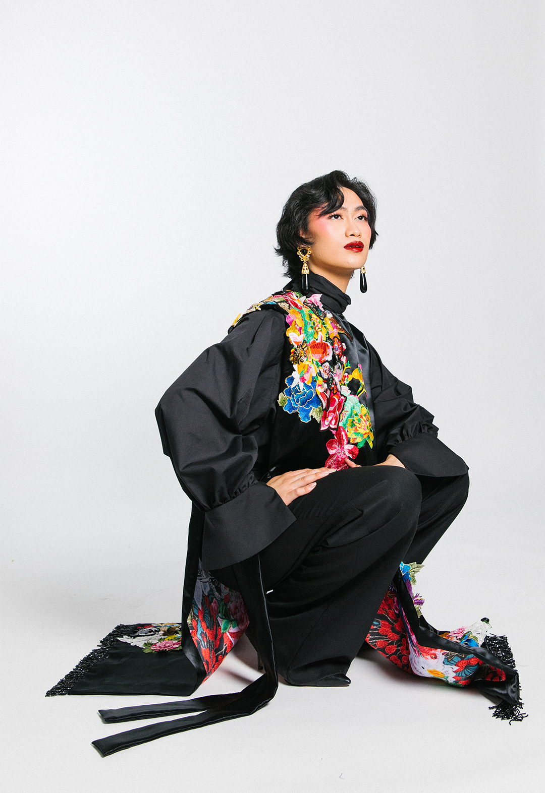 The model is crouched in black wide-legged wool pants with a black cotton poplin tie-neck blouse under a black satin poncho with collaged clusters and a printed lining.