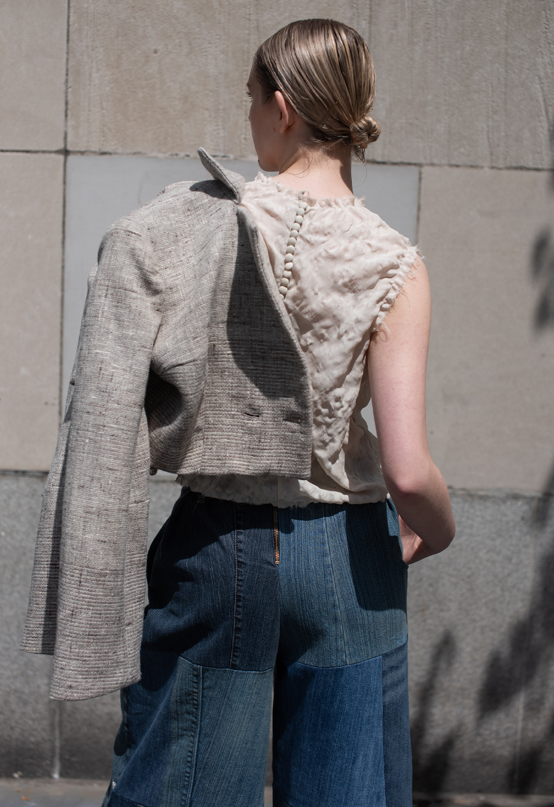 The photo is a back-view image of a girl displaying closure details of beige self-covered buttons on the hand-manipulated blouse and a brass metal zipper that is applied at the back of the denim pants. The wool jacket is placed on her shoulder as one of her hands is placed inside the pocket of the pants. Her head is turned slightly to the side. The background has multicolored gray and beige concrete panels. 