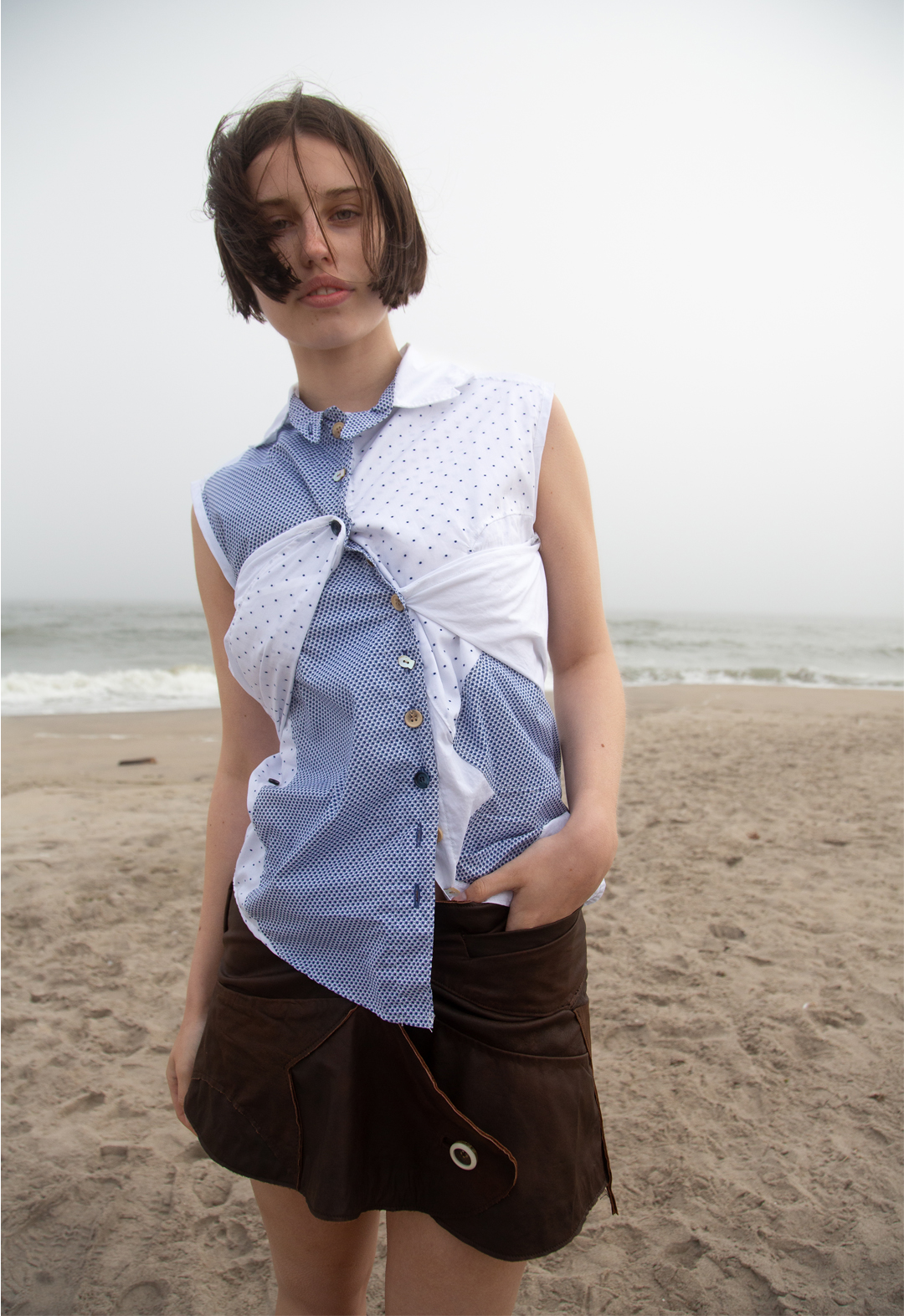 A model wears a blue-and-white muti-print button-up top that detaches from the shoulders and buttons into the center front buttons. Each button is either a brown or blue wooden button or a fish-shaped mother-of-pearl button. The top is paired with a wrap-around dark-brown leather skirt constructed from fish-shaped pattern pieces. The skirt has both a front and side welt pocket and two large, clear vintage buttons that close the skirt in the front.  