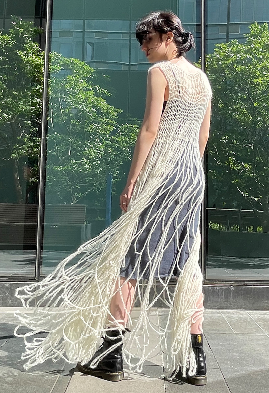 Back view of a model wearing a hand crocheted white lace suri alpaca dress. The model is wearing combat boots and there is a glass building in the background, with reflection of trees.