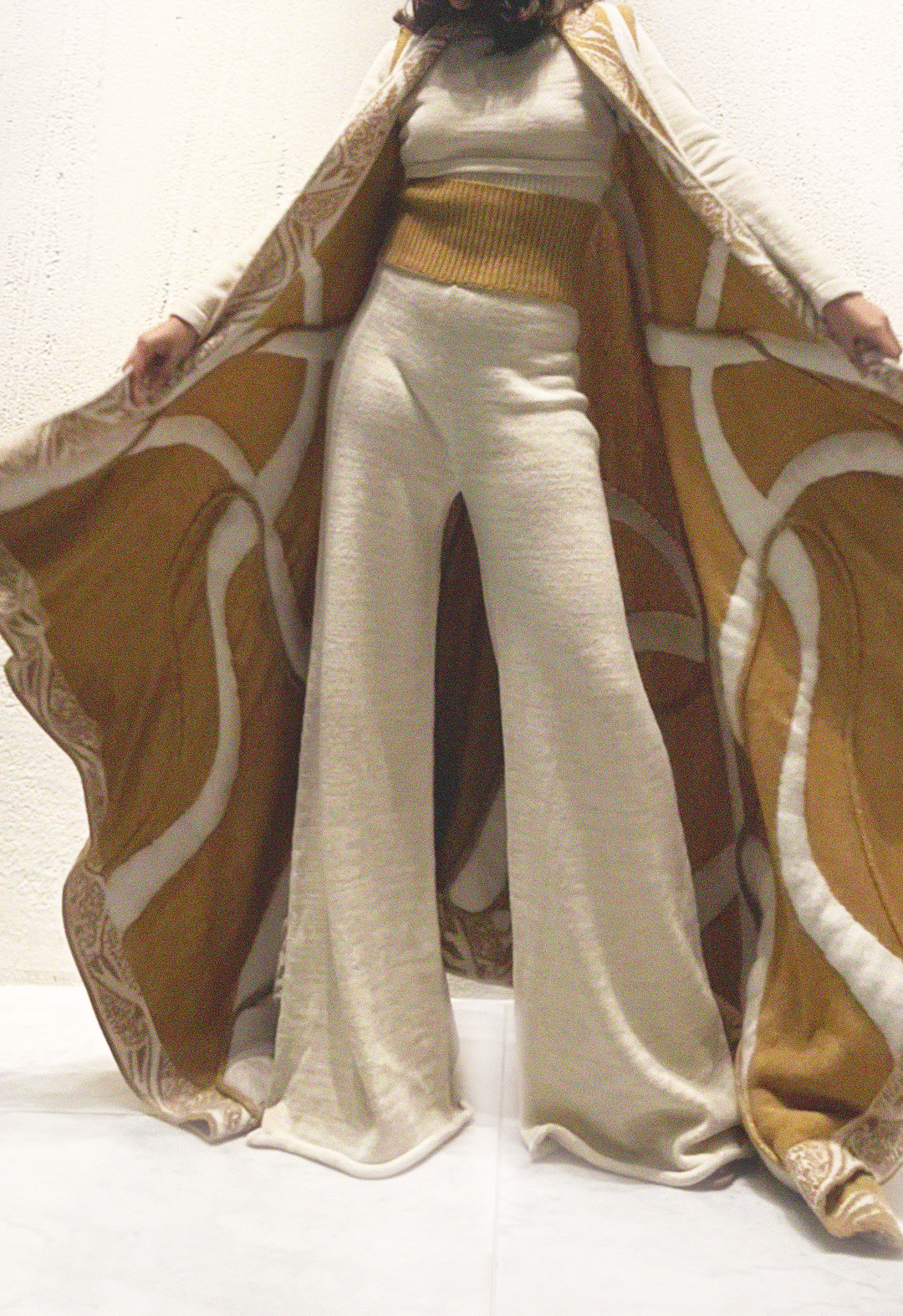 The model is wearing a two-color jacquard pattern cardigan that has a flowing line pattern all over, which is stuffed to make it look more sculptural. The cardigan is paired with matching white full-sleeve top and white legged pants with a double-rib waistband. She is holding the cape wide open with her hands, which allows us to see the inside of the reversible cape and a full view of the pants and top. The background is white.