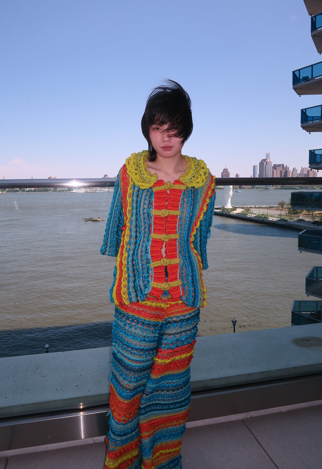 Front view of a model wearing a knit suit. The yarns alternate randomly between red, blue, and yellow. There is a river in the background.