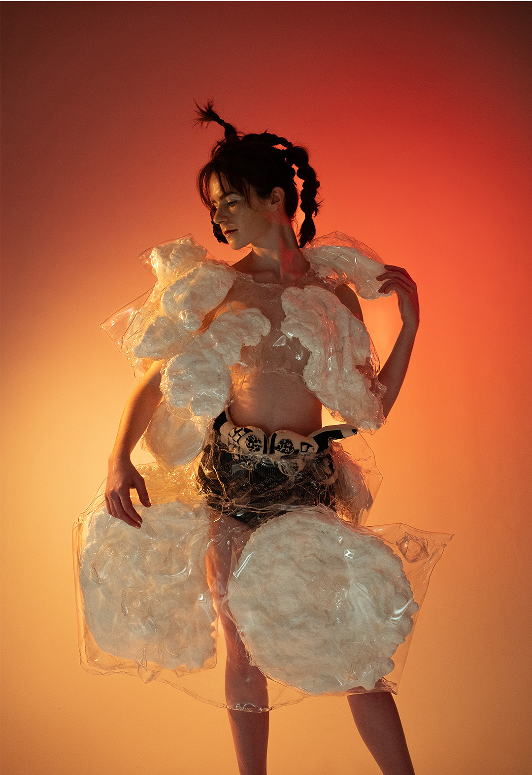 Front view of a model wearing a worbla structural piece with installation foam encased. She also wears panties with dolls under the worbla. The background is a top to bottom gradient from dark orange to yellow. 