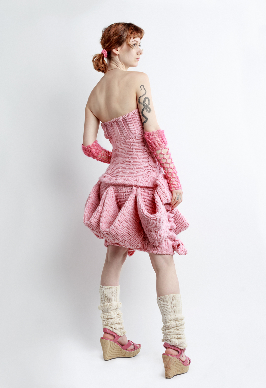 Photo of a girl posing in a pink sleeveless magical girl mini dress, looking to the side. The dress is hand knit with baby pink pink wool. The dress features big bows and big pockets circling the skirt of the dress. The dress, and girl, are posed at a three-quarter back view to the camera. The girl is wearing crochet fishnet gloves, heels, leg warmers, pigtails, and a heart necklace. The girl has her far arm raised slightly.