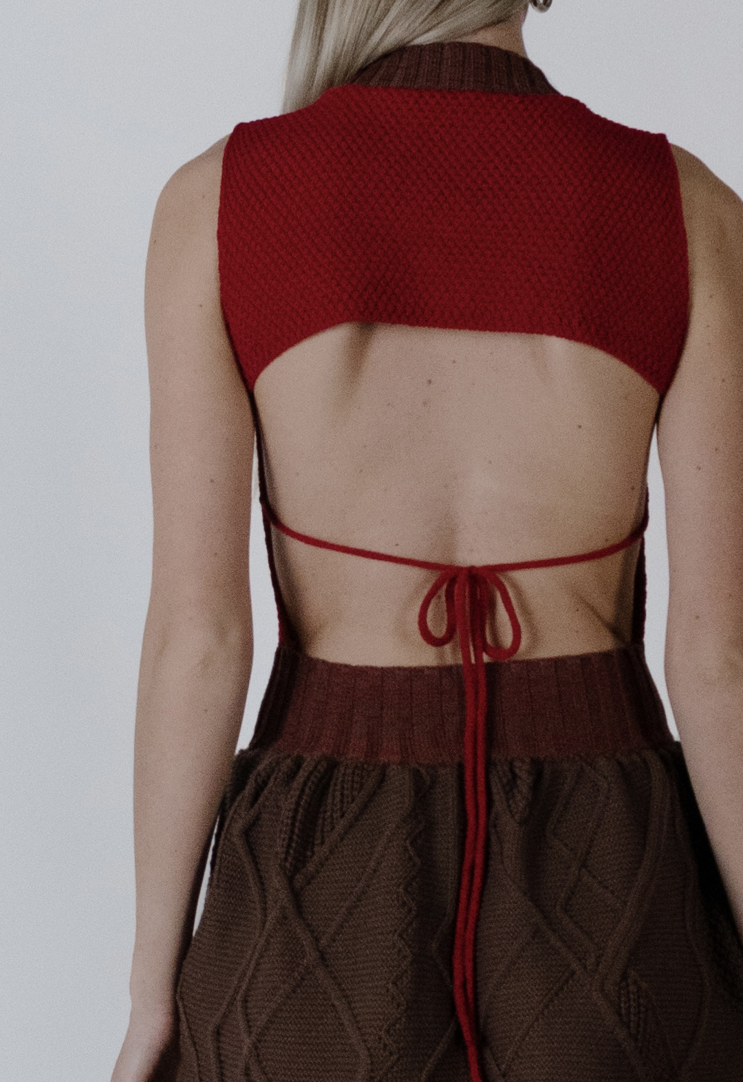 In this photo, you see the model's back with her arms to the side. The tight-fitting top of the dress with moss stitching and the lower back are shown, with a bow across the model's back. You can see the top portion of the Aran cable skirt and the heather red rib at the waist. The background is white.