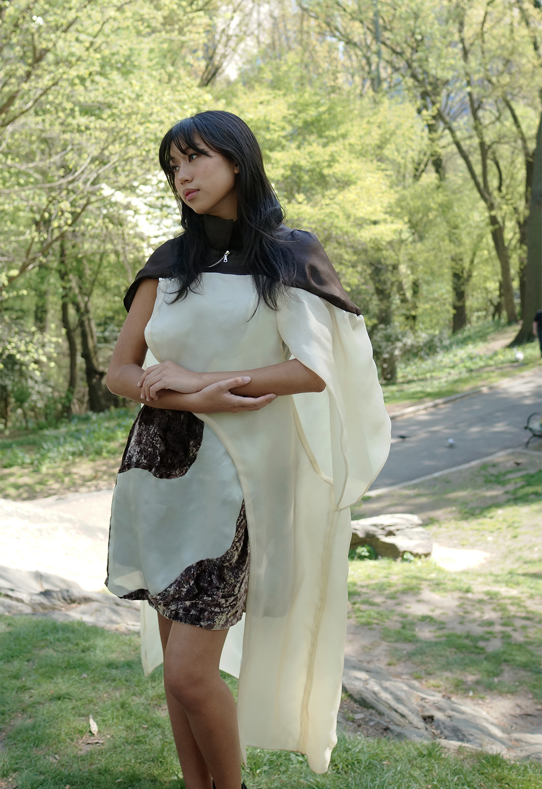Front view of a model wearing a brown and white organza cape and paneled velvet dress. There are trees in the background.