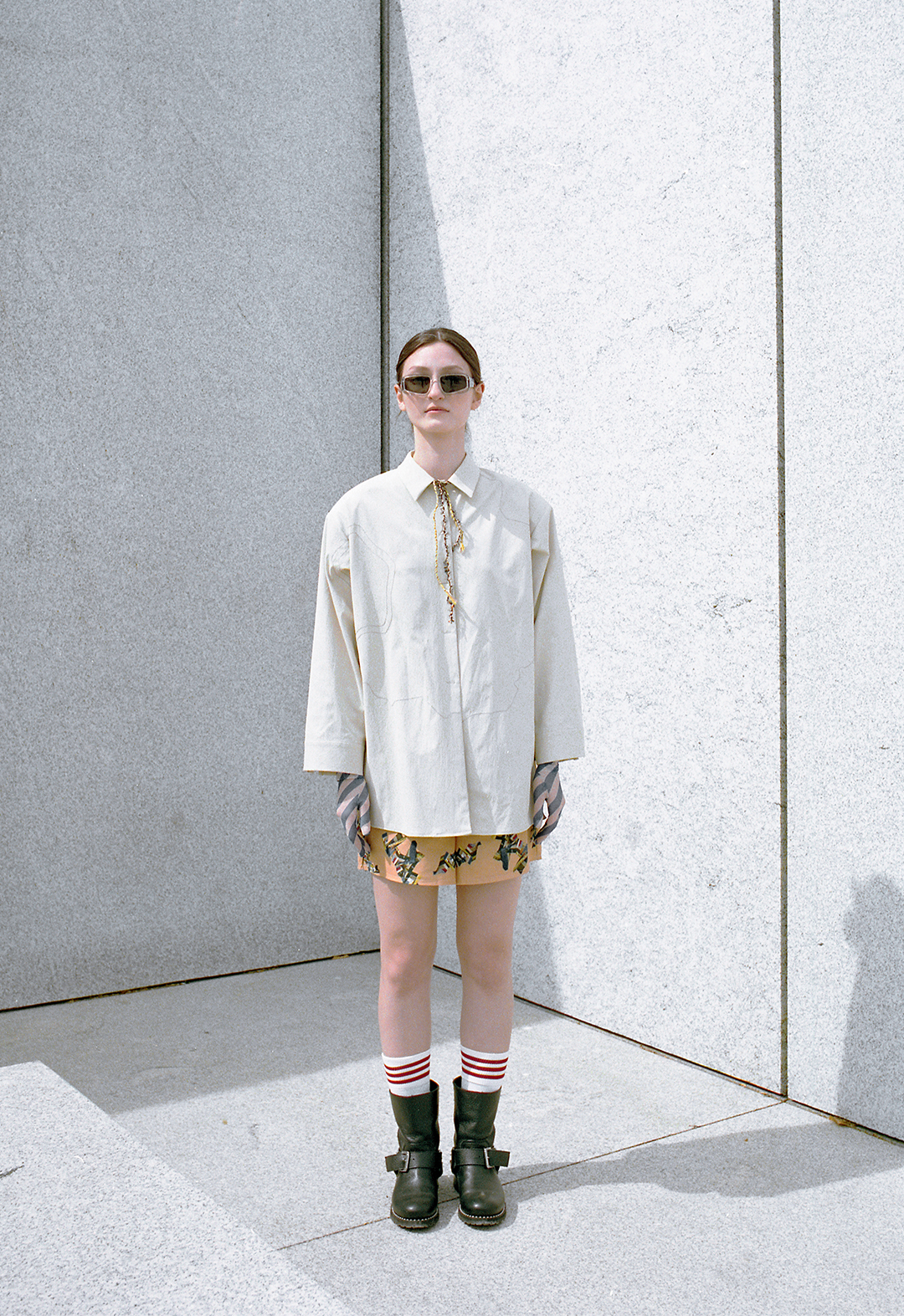 This is a green, embroidered, oversized shirt, worn with patchwork digital printed shorts. It is accessorized with sunglasses, red-striped socks, and black boots. The model is posed in front of a gray wall.