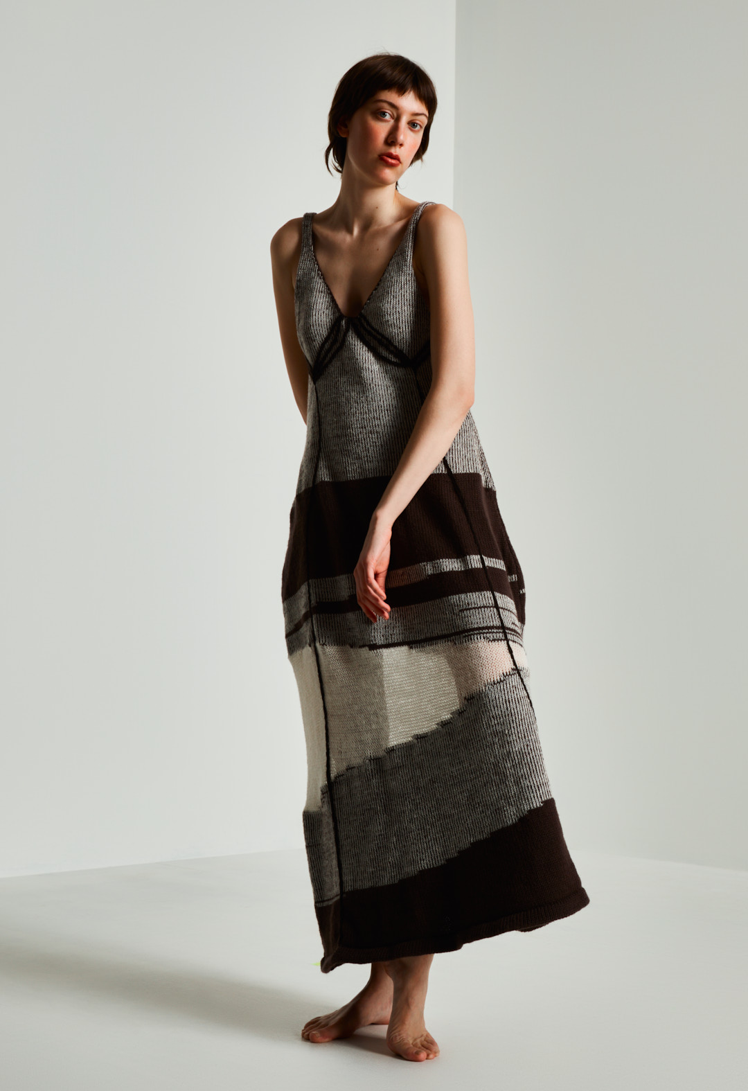 The photo features a model in a brown-and-cream intarsia midi dress with translucent detail, linear embroidery, and plaiting, made of organically certified wool. 