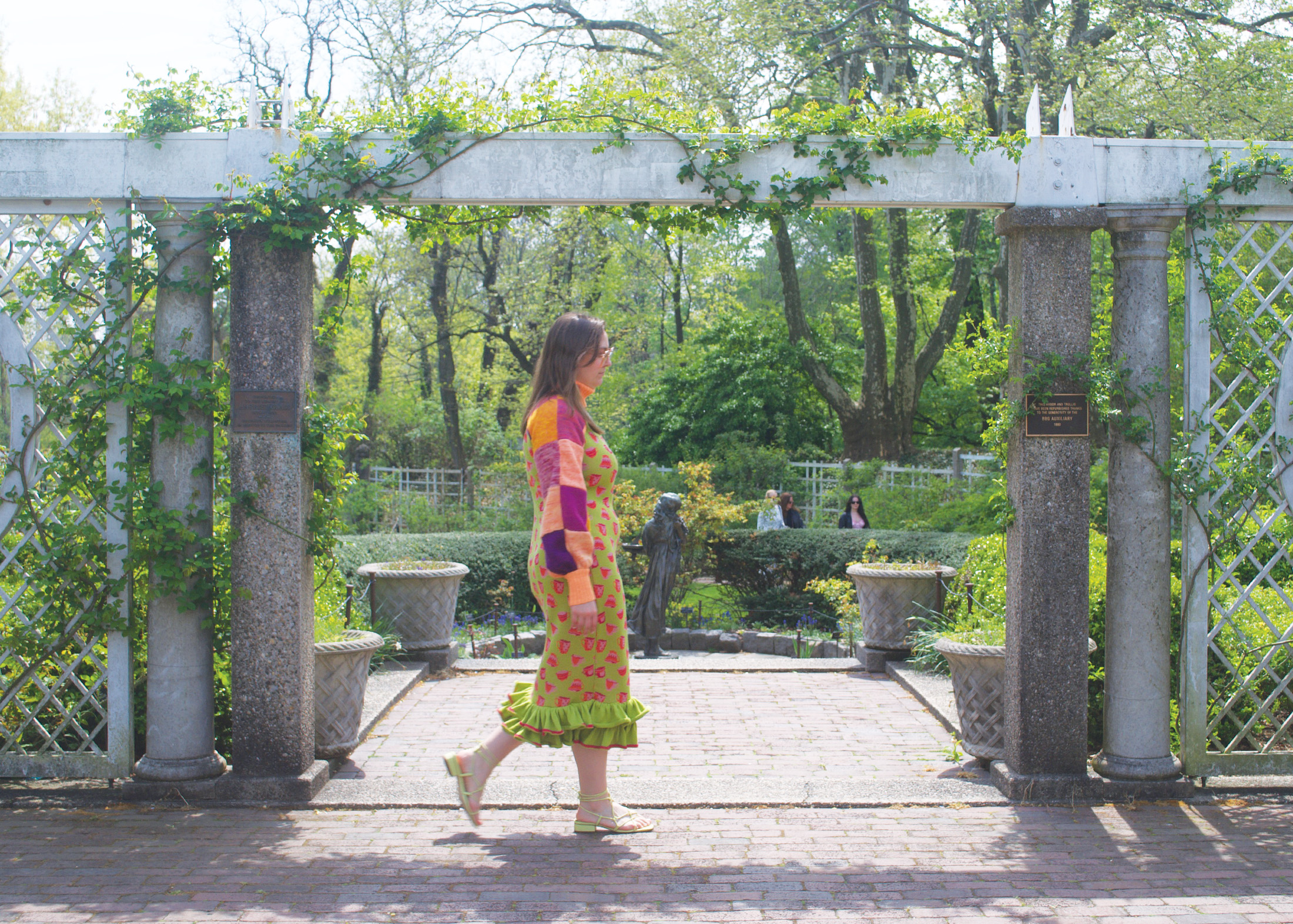 Photo of a model wearing a green, teacup-motif dress with a ruffle hem and a purple-and-orange patchwork turtleneck shrug. She is walking across the frame in front of a garden.
