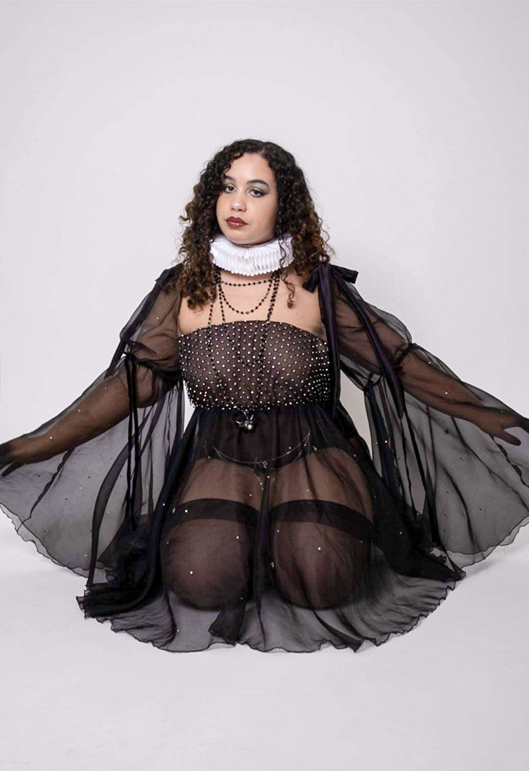 The model is sitting kneeling in a black organza babydoll dress with rhinestone netting, over a black mesh panty  with chains.