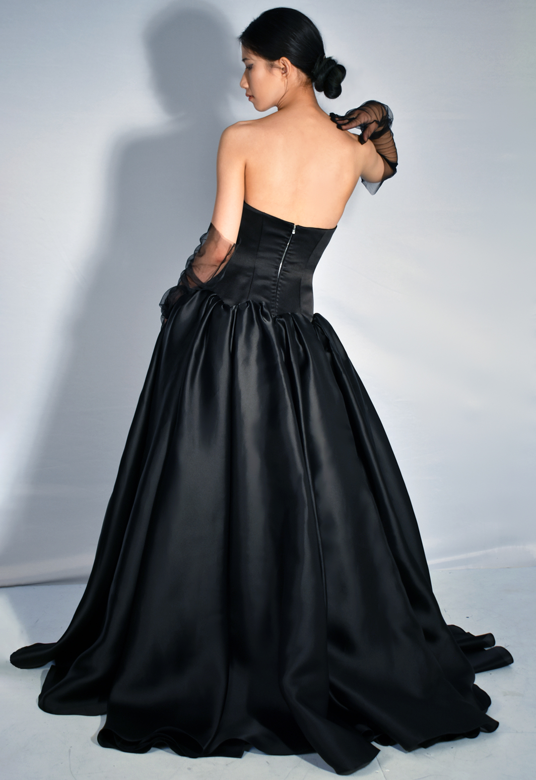 This black gown is made from 100 percent silk. The photo shows a back view of a woman wearing a black floor-length silk gown that has a drop-waist duchesse satin bodice with cone-like satin-faced organza skirt panels gathered along the drop-waist, pooling onto the floor. The back of the gown has a silver metal zipper. The model is wearing black tulle gloves that cover three-quarters of her arms. Her head is turned to the side, and her right hand is sitting on her shoulder. The background is white.