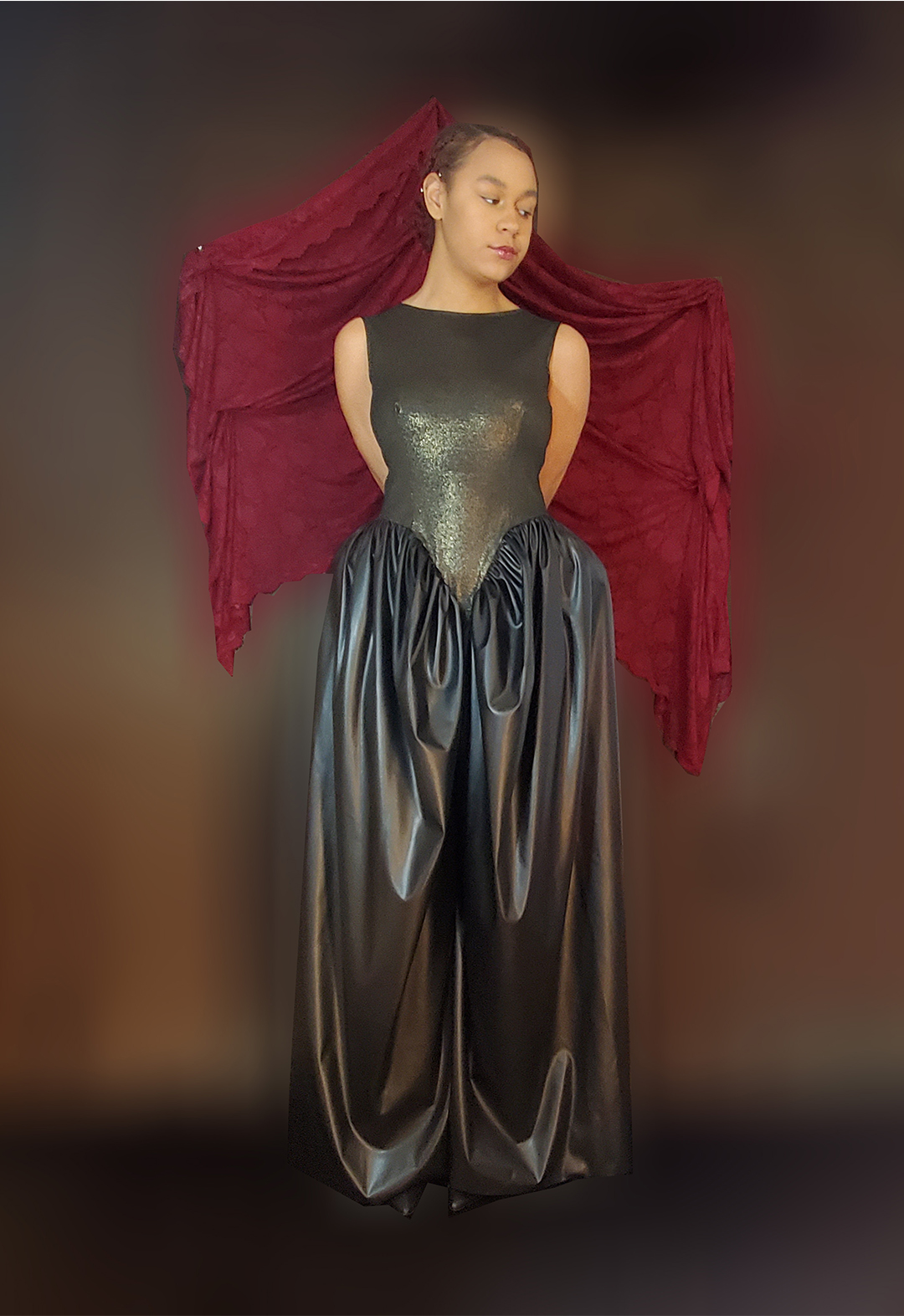 A model wearing a sleeveless jumpsuit that has a knit bodice and leather pants. The pants have gathering at the hips to resemble late 18th-century French dresses. The model is posed with her hands behind her back while looking down. The background is moody.