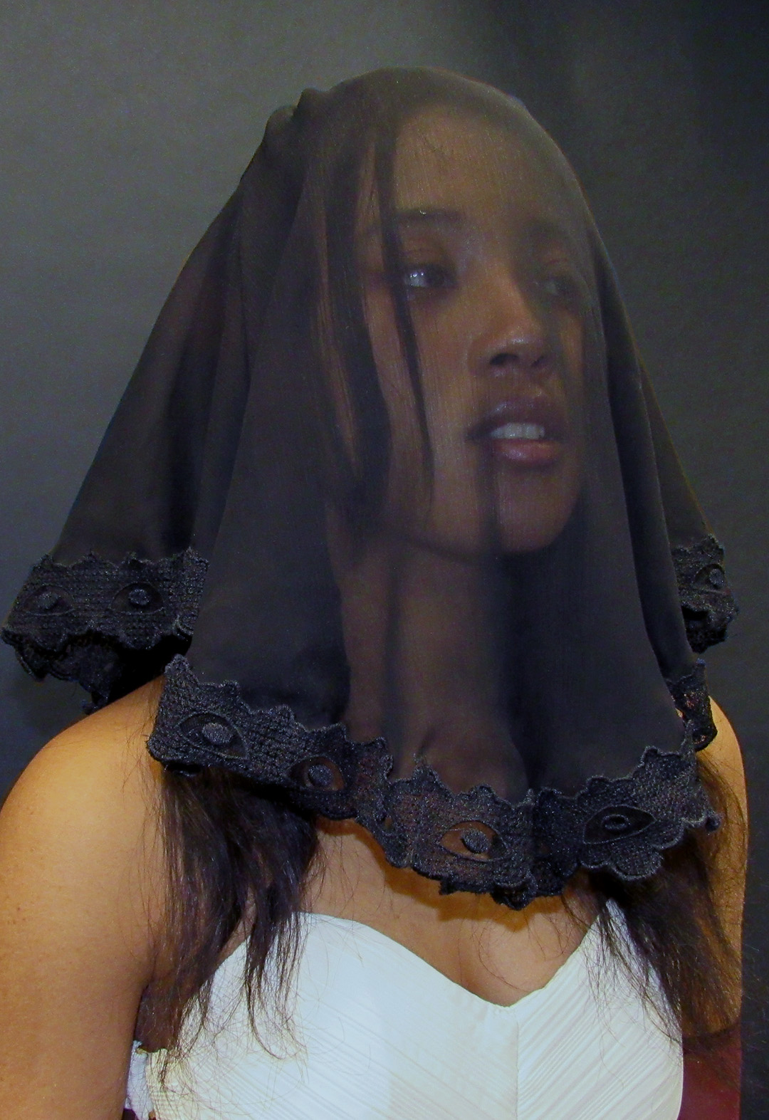 A close-up shot of a model's face, looking away from the camera. Her face is covered with a black, draped, embroidered veil. Her upper arms are exposed, and she wears a textured, ivory bustier.