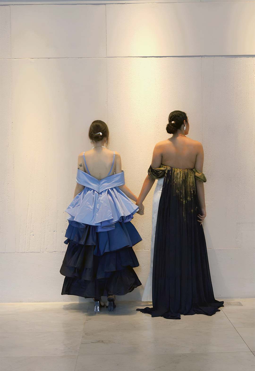 Back view of two models. The model on the left is wearing the off-the-shoulder tiered silk taffeta gown in multiple shades of blue. The model on the right is wearing the pearl-white satin gown with silk navy blue and hand-painted gold chiffon train. 