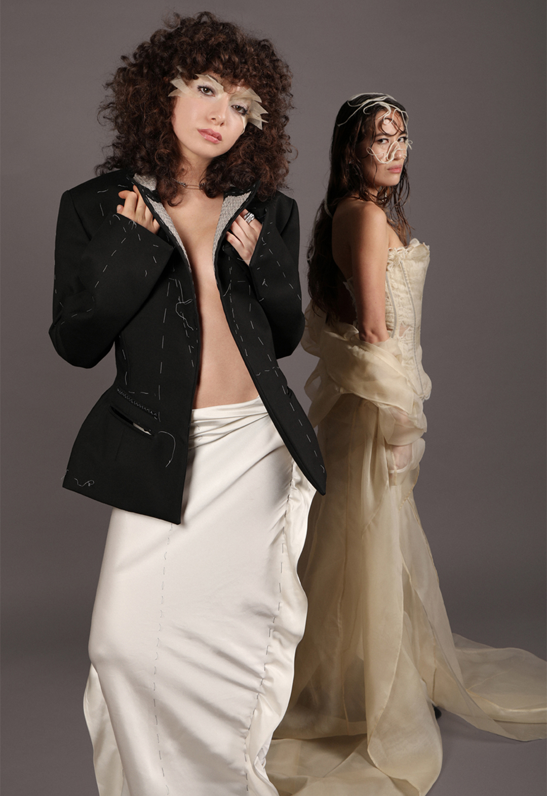Two models presenting the two looks. The model closest to the camera is wearing a back tailored jacket and a bias-cut ivory skirt. Behind her and farther from the camera is another model wearing a sheer corset top with a sheer draped skirt/jacket.