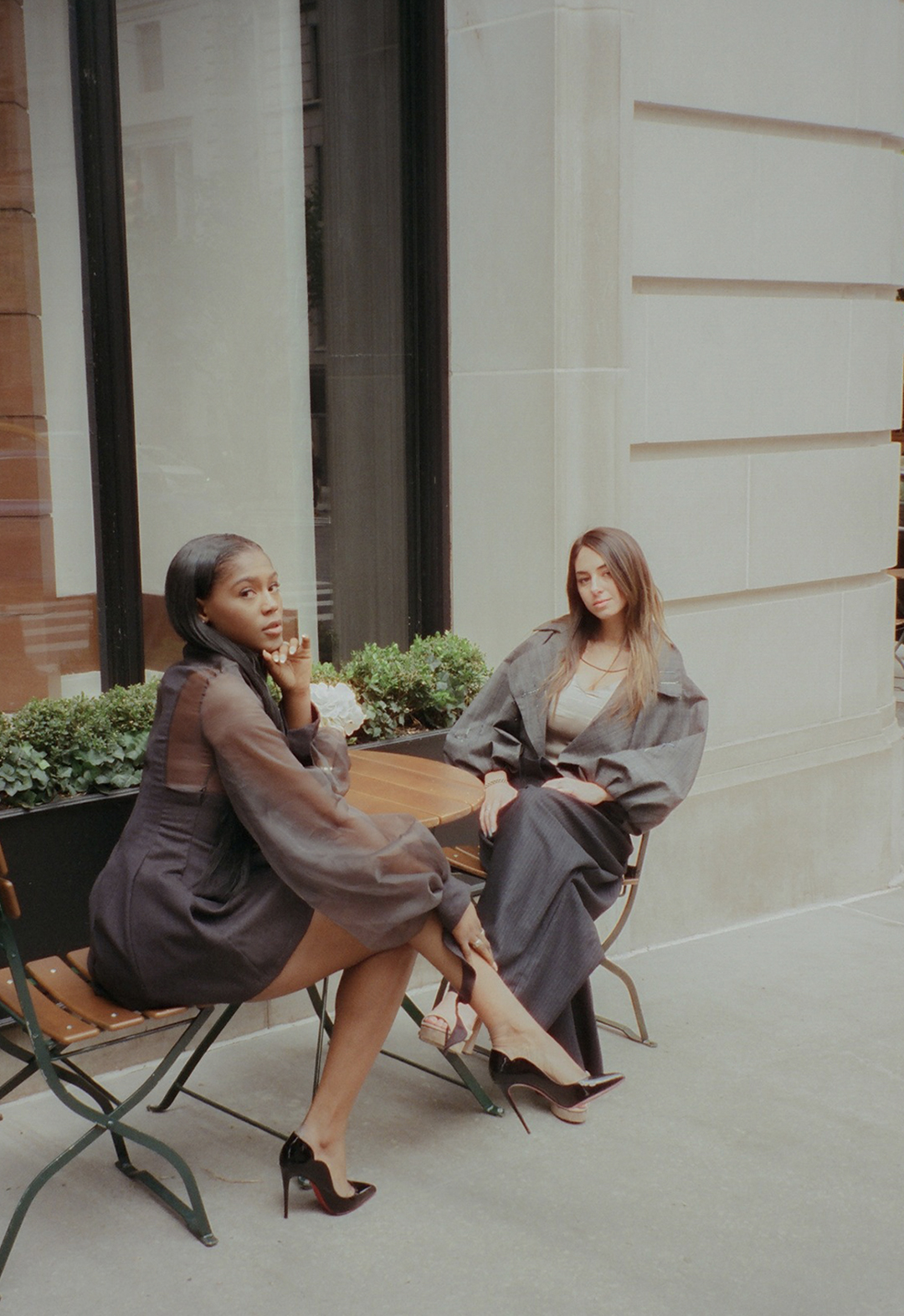 Two models sitting outside at a small table in an urban setting. The model on the left is looking at the camera. She is wearing a dress with heels. The model on the right is wearing pants and a bomber blazer.
