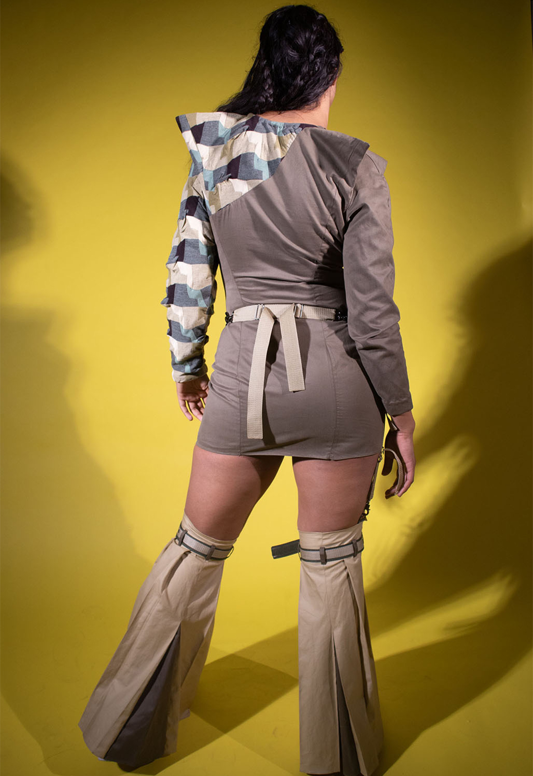 A young woman stands in a studio with a vivid yellow background, highlighting the back of her innovative attire. She dons a nightingale brown dress complemented by a turquoise color-blocked sleeve with pointy shoulders and pairs it with khaki skirt-style leg warmers for a daring and distinctive look.