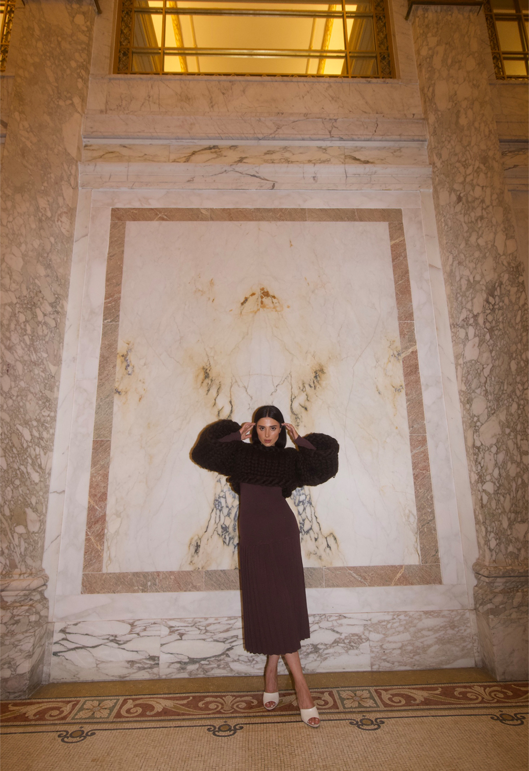 A model standing in front of a white and brown marble wall, wearing a brown dress layered with chunky hand knitted shrug.