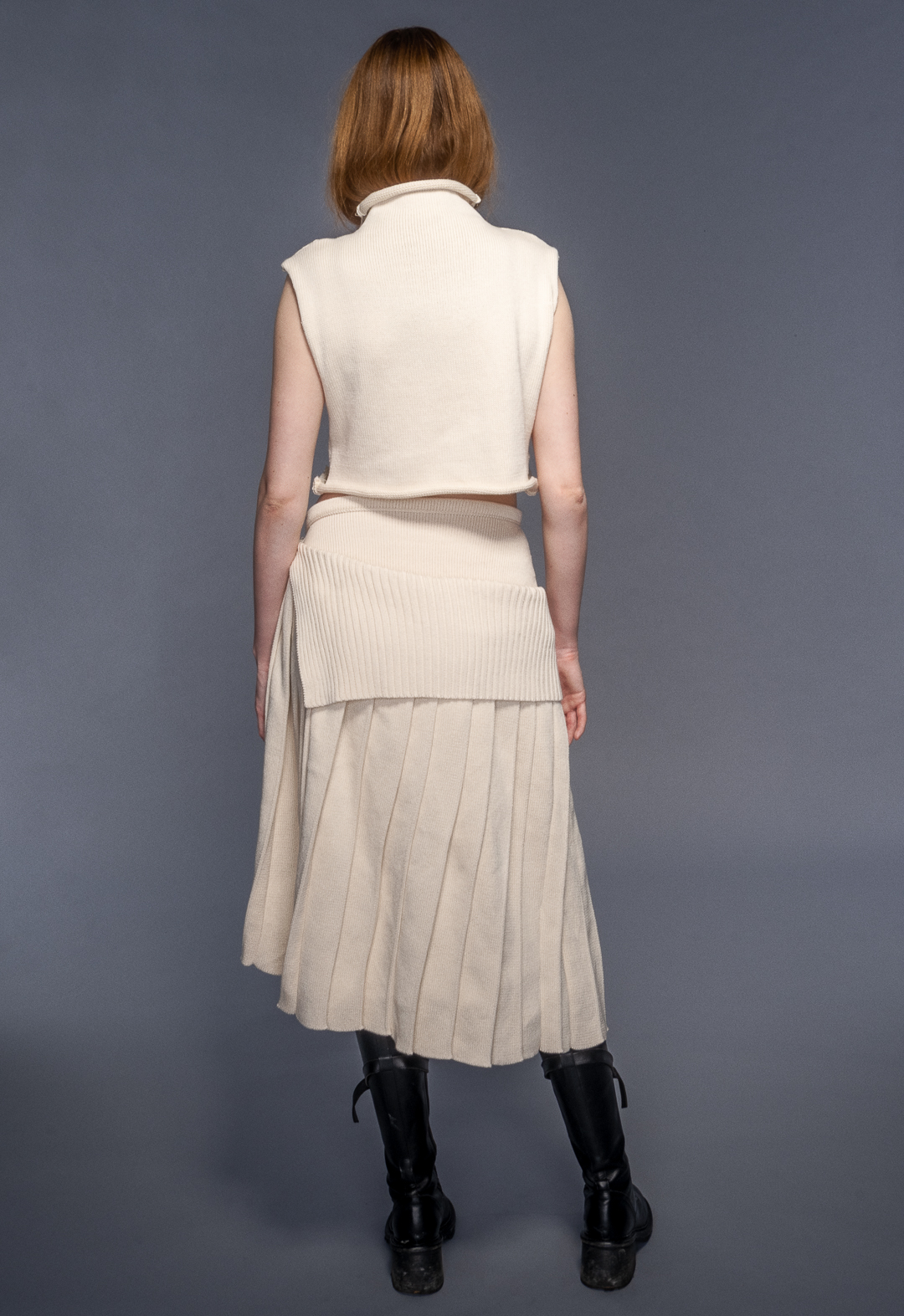 Model wears funnel neck top with misshaped intarsia and irregular pleated skirt in organic cotton. Her hair is tucked in the sweater.