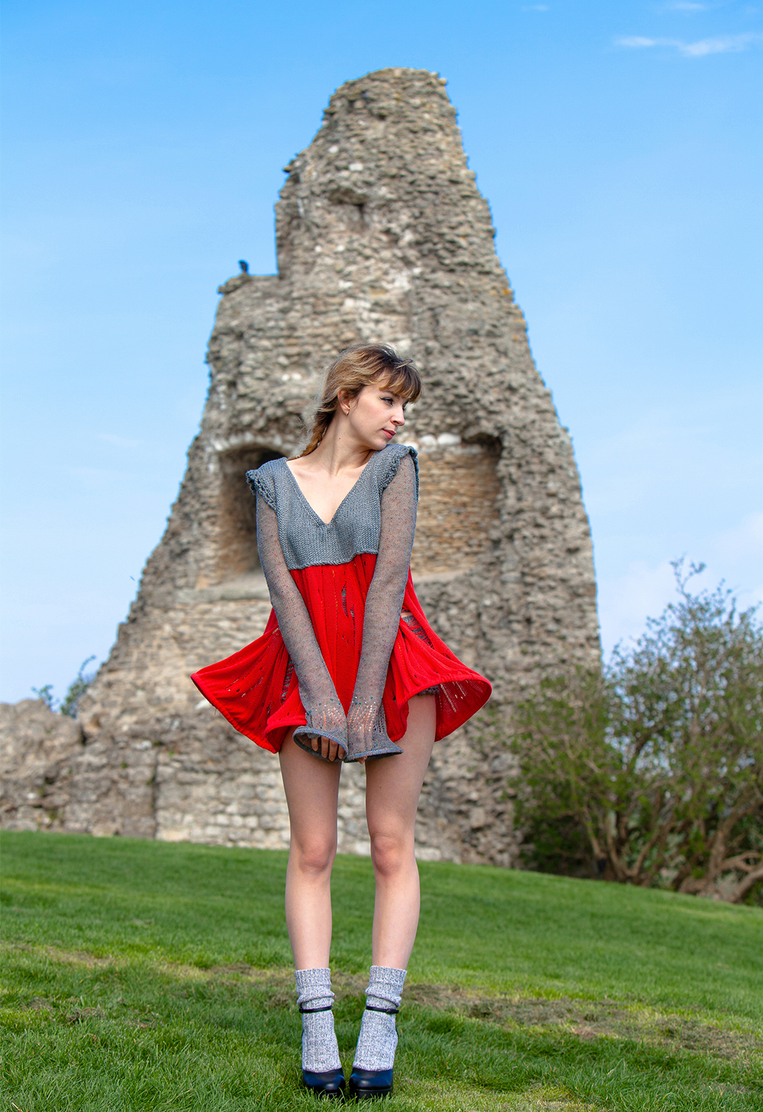  A model is posing in front of a castle in ruins with her hands in front of her. She is wearing a gray-and-red knitted fairy-tale dress, gray knitted briefs with a crochet edge, and a sheer lace top with bead accents.