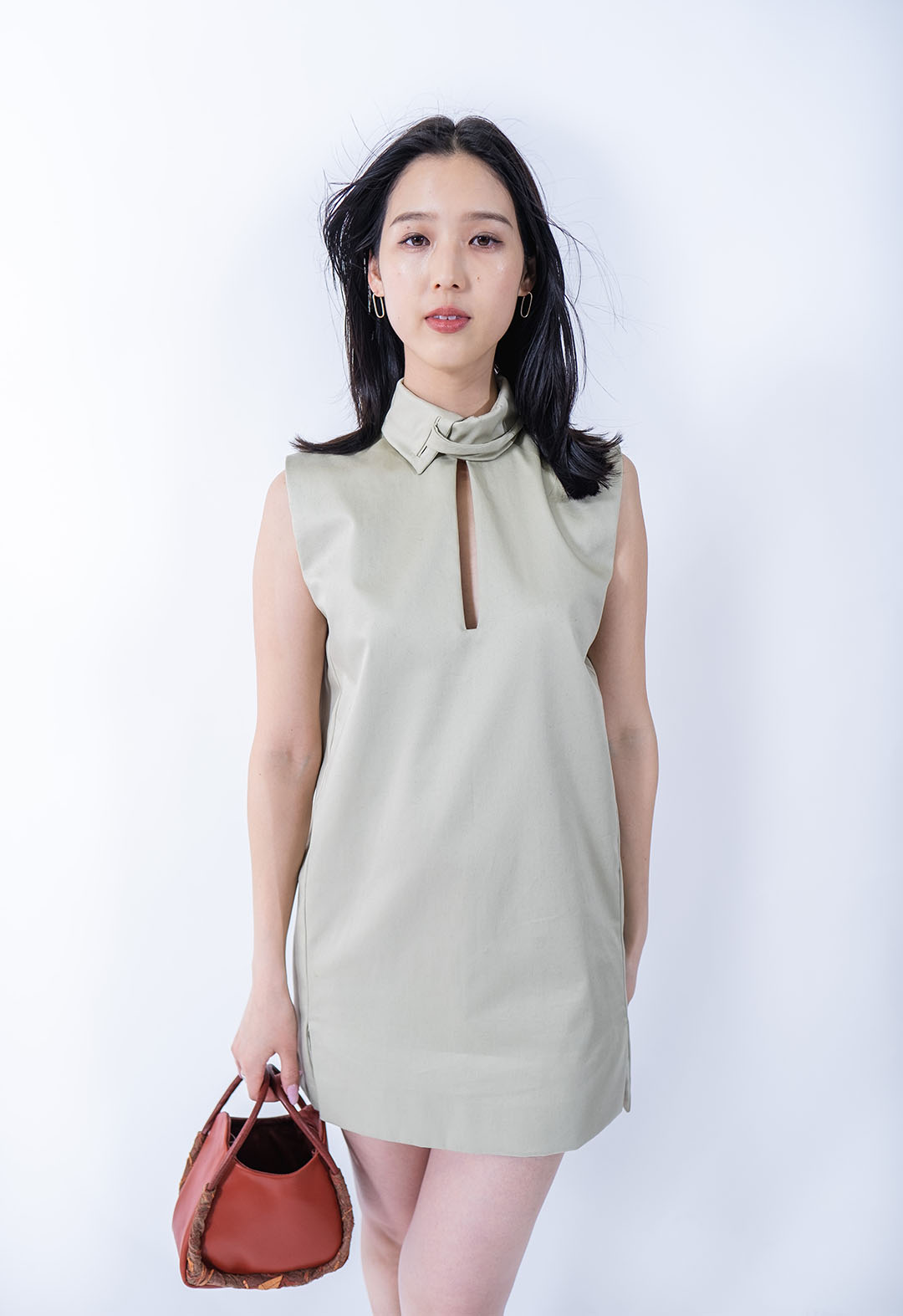 This pale, light-green mini dress has a skinny rectangular cutout from the neck to the bust. There is a stand collar attached to the neckline that pulls through itself via welt openings to tie into a bow at the side of the neck. The model is standing in front of a white background and looking straight into the camera with one hand on her hip.  
