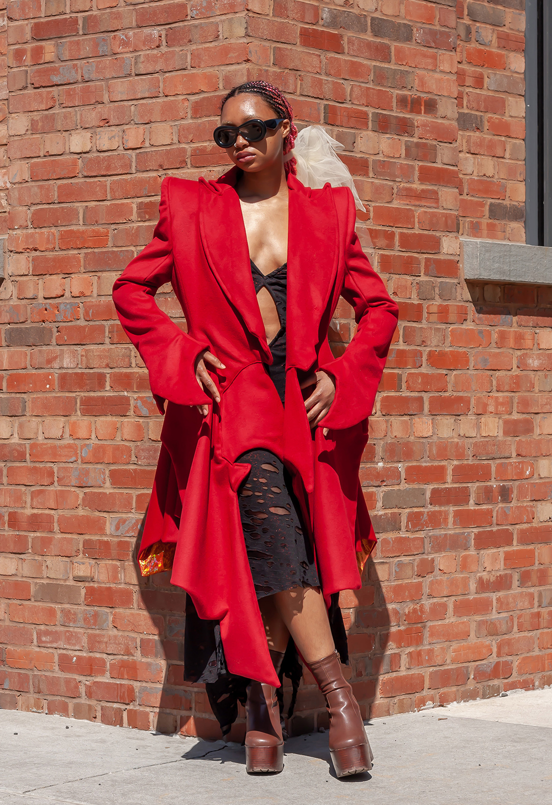 Front view of Traci posing in the Burgeon Coat. She has her hands on her hips and her legs are crossed. She wears black sunglasses and is standing in front of a brick wall. 