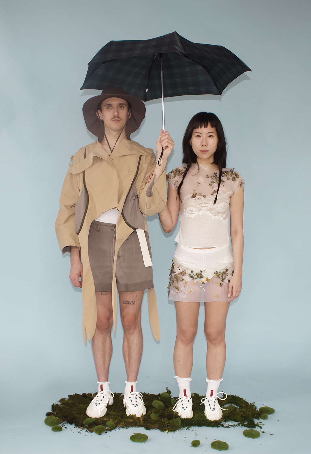 The couple share an umbrella while standing next to each other. The male model holds the umbrella while the female model rests her right hand on his left arm.