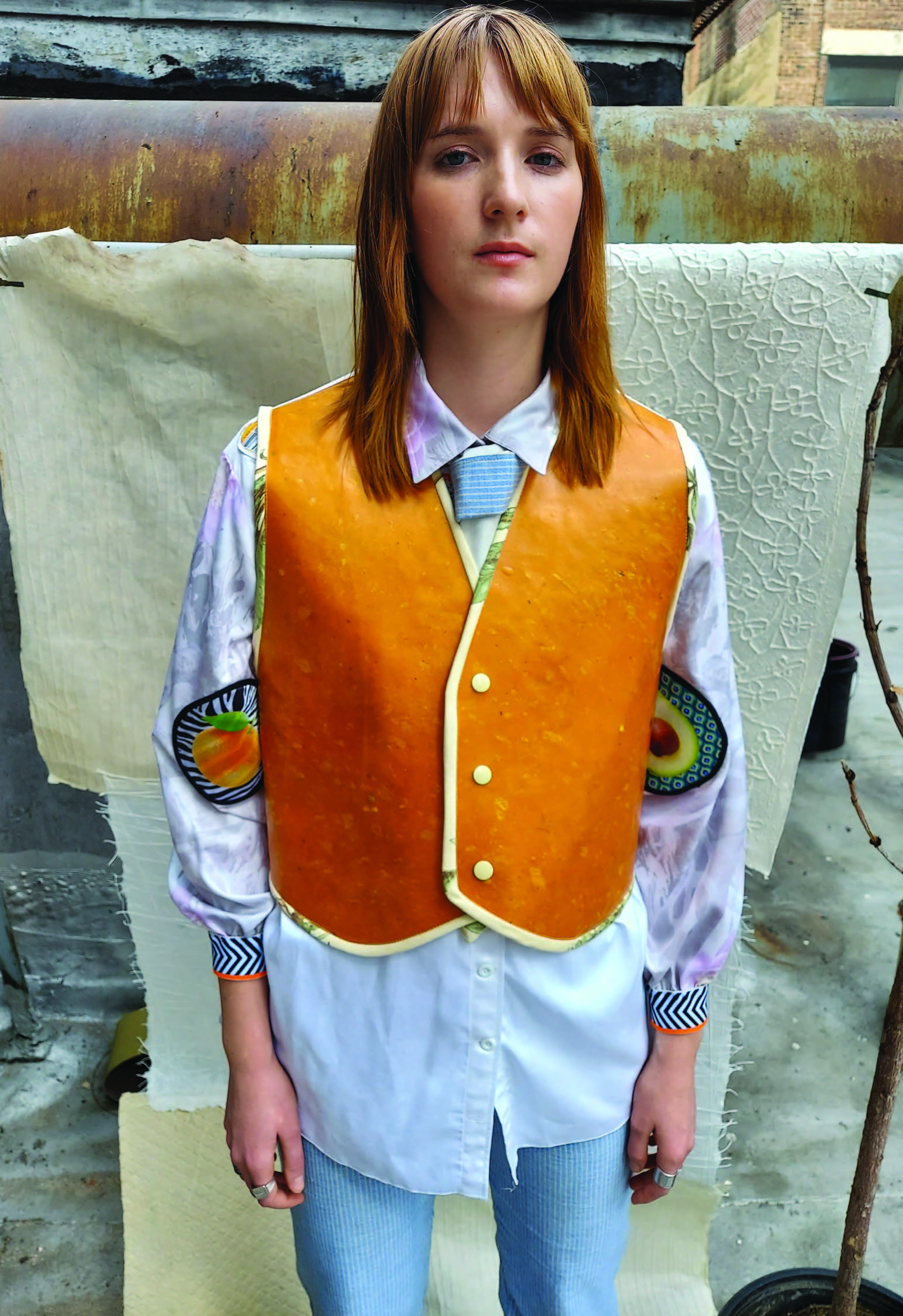 The model wears a button-down dress shirt made from the eucalyptus plant, with one-to-three-inch fruit sticker patches arbitrarily placed on the torso. A faded heat transfer print is shadowing other patches; larger fruit patches are placed at the elbows. A patchwork tie is in a full Windsor knot around the neck. The vest is made out of a vegan leather from the lotus plant, sourced from Cambodia. There are three snaps at center front, and an adjustable sizing tie at the back. The back also has a heat transfer print of fruits. The light-blue striped trousers are made of 95 percent organic cotton and 5 percent lotus. They have a six-button fly, a hammer loop, and four pockets.    