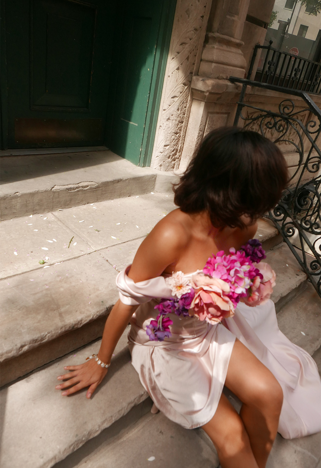 3/4 top view of a model sitting on stairs, wearing a baby pink off-shoulder dress with draped cowl and side train with flowers on the neckline.